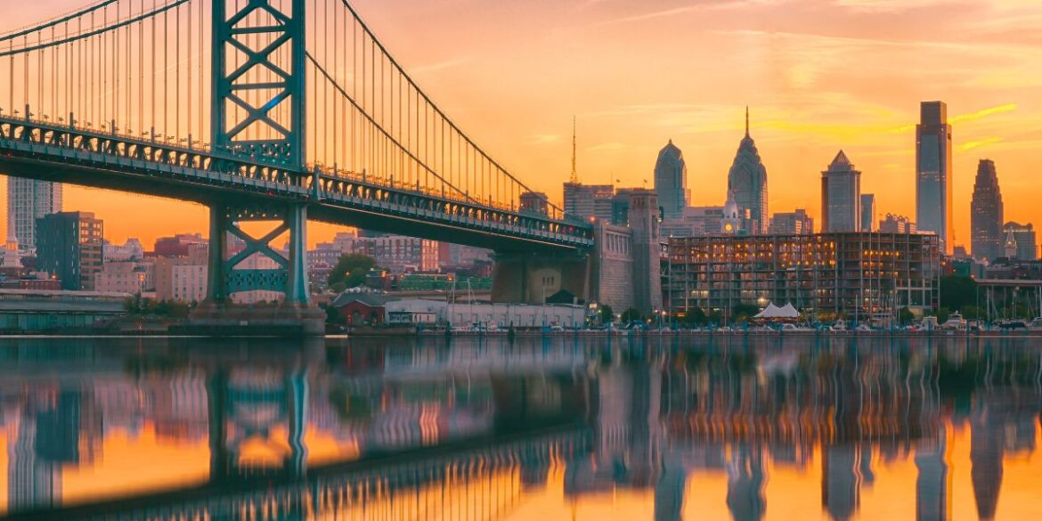 A romantic view of the Philadelphia skyline, water, and bridge at sunset.
