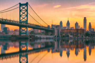 A romantic view of the Philadelphia skyline, water, and bridge at sunset.