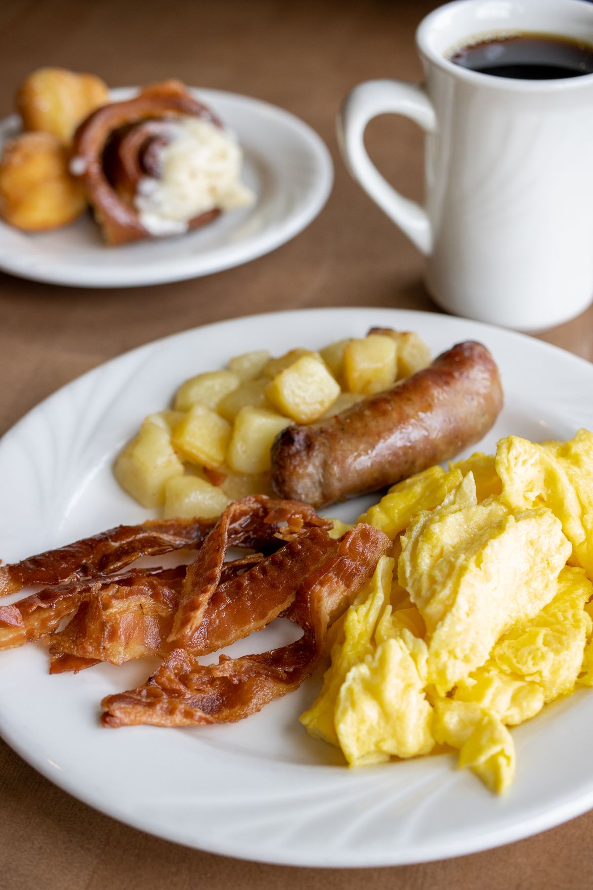 A well-balanced breakfast plate featuring scrambled eggs, crispy bacon strips, a sausage link, diced potatoes, and a cinnamon roll. A white mug of coffee completes the meal.