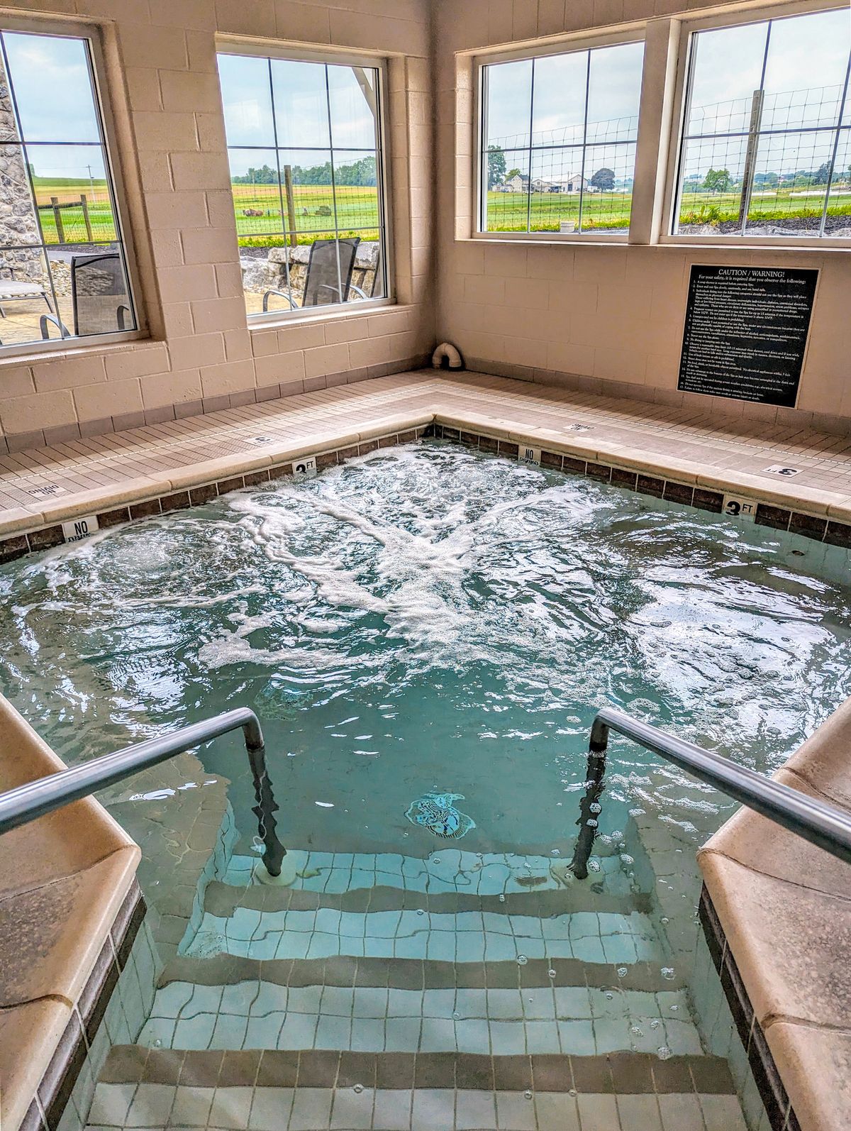 Interior view of a jacuzzi at Amish View Inn with agitated water. The jacuzzi is surrounded by beige tiles and has steps leading down into it. Large windows offer a view of the outside, showcasing green fields and a fence. A black plaque with white text is visible on the wall.