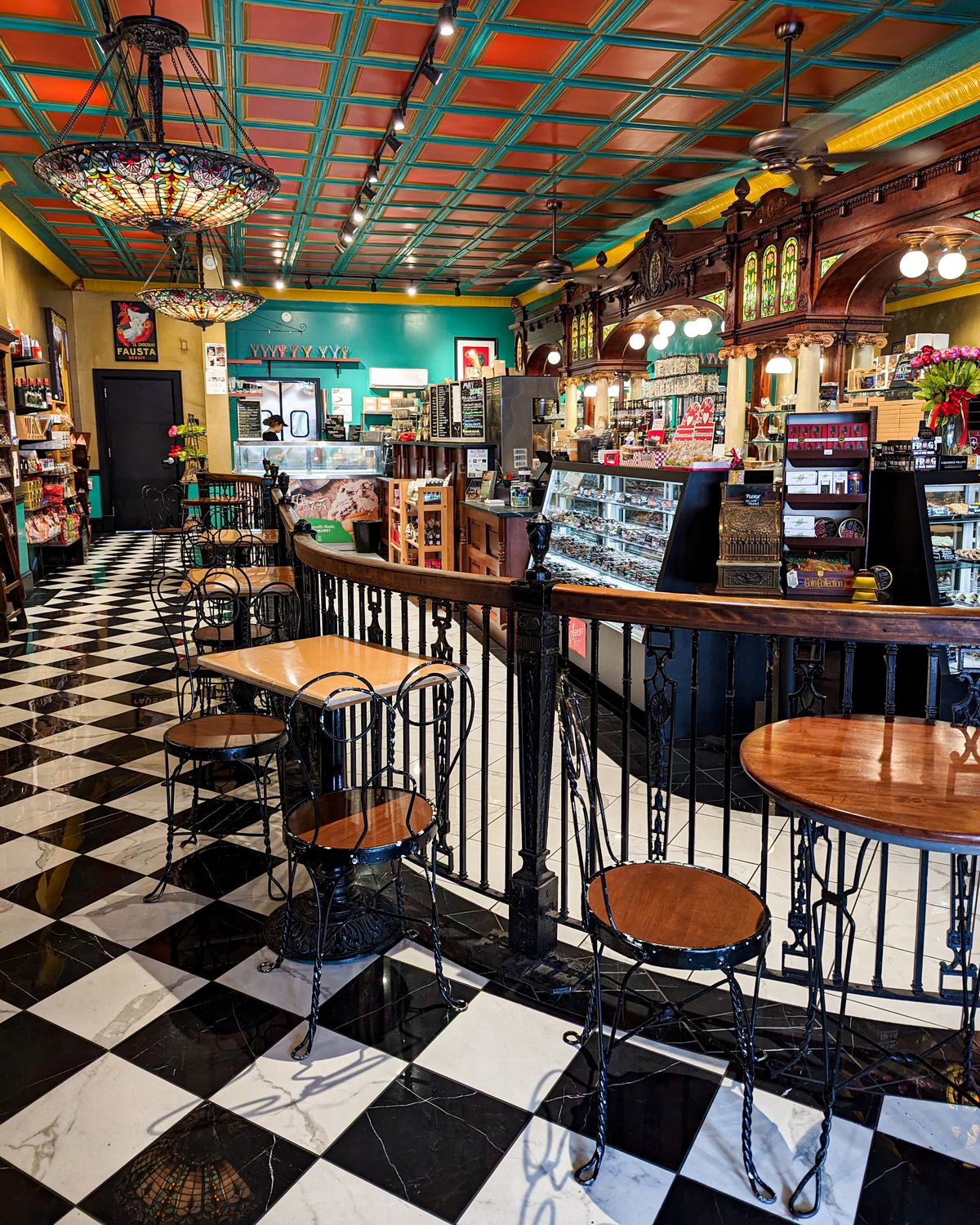The image shows an interior view of a charming vintage-style café. The café features a beautifully intricate ceiling with a colorful, geometric pattern, and two elegant stained-glass chandeliers hanging from above. The floor is a classic black and white checkered design. The seating area includes black wrought iron chairs and small wooden tables, adding to the old-fashioned aesthetic. The counter area displays a variety of sweets, pastries, and other treats, with antique wooden cabinets and stained-glass panels enhancing the nostalgic atmosphere. A touch of modernity is seen with the presence of various products and menus behind the counter.