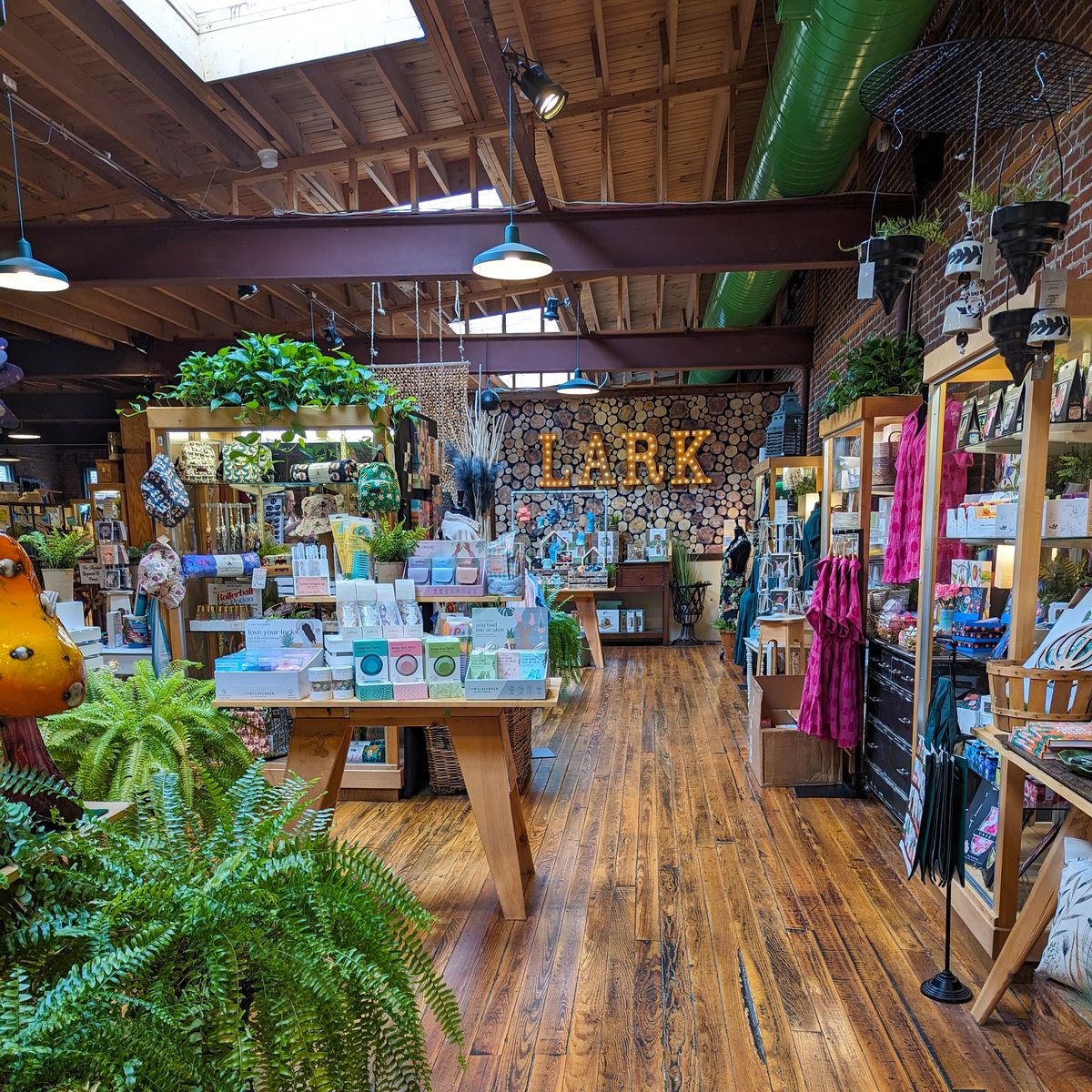 The interior of retail store "LARK". The store has a rustic, wooden interior with exposed beams on the ceiling. The shop features a wide variety of merchandise displayed on wooden tables and shelves, including plants, figurines, jewelry, candles, soaps, and other decorative items. A large illuminated "LARK" sign is visible on the back wall. The overall atmosphere seems cozy and inviting, with warm lighting and an abundance of greenery from the many potted plants throughout the store. The wooden floors and natural materials used in the displays contribute to the shop's charming, bohemian aesthetic.
