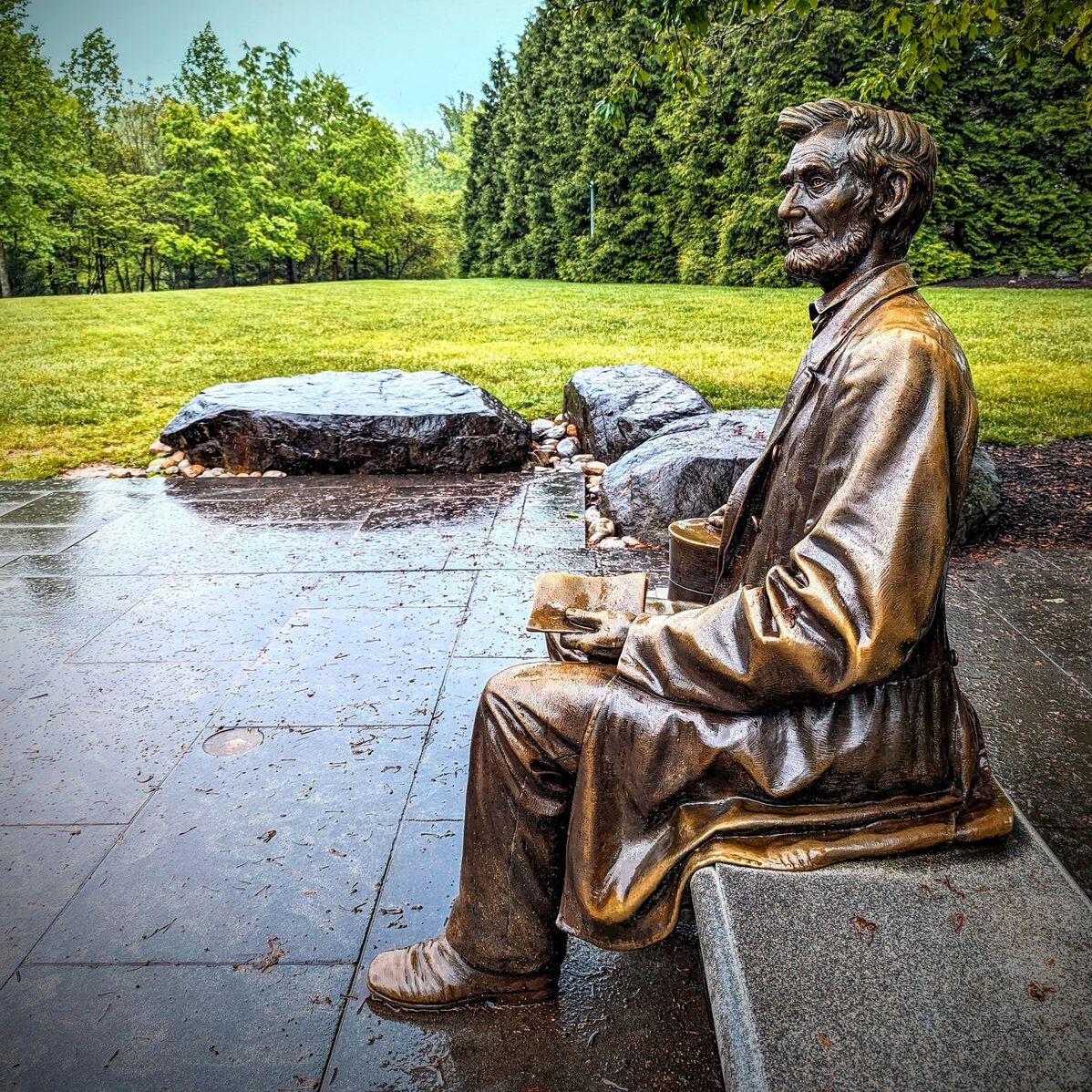 A bronze statue of Abraham Lincoln seated on a stone bench, holding a book on his lap, located in a serene outdoor setting. The statue is surrounded by lush green grass and trees, with large rocks positioned nearby on a wet stone pathway, adding to the tranquil ambiance.