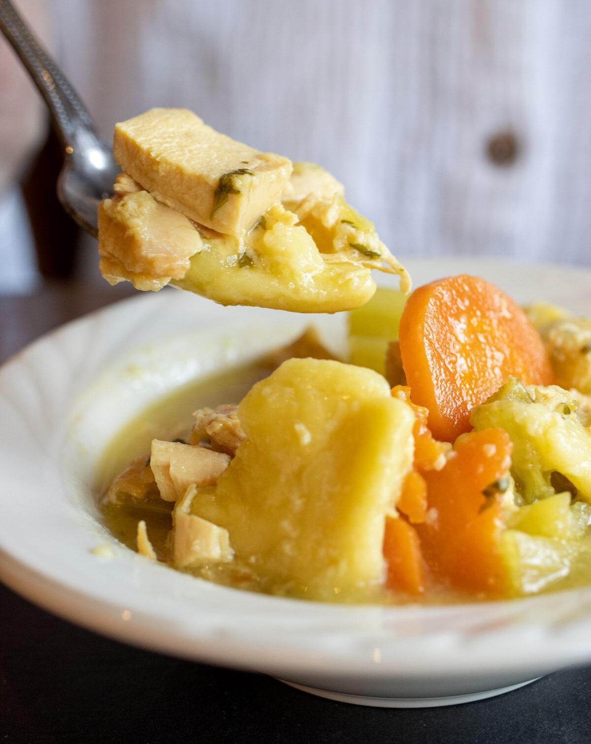 A bowl of chicken pot pie with chunks of chicken, carrots, and potatoes visible in a creamy broth. A spoonful is being lifted from the bowl.