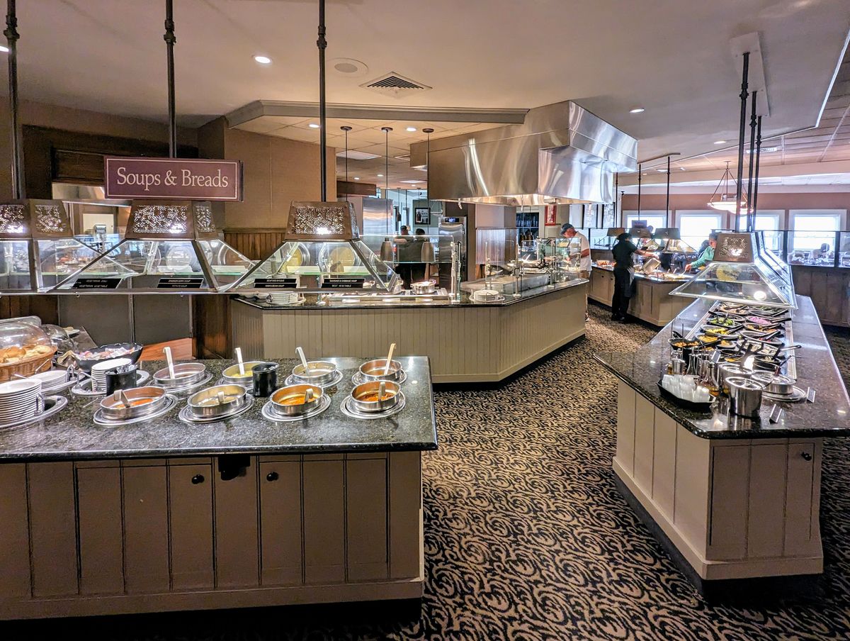 Interior view of Miller's Smorgasbord showing multiple food stations, including a "Soups & Breads" area. The space has carpeted floors and pendant lighting. 