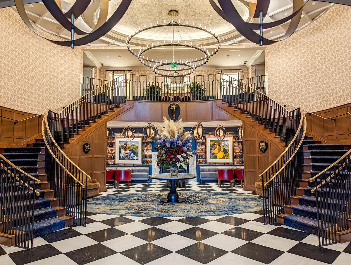 The lobby of The Grand Lodge at Nemacolin. Two staircases, marble floors, seating areas, and a chandelier. 