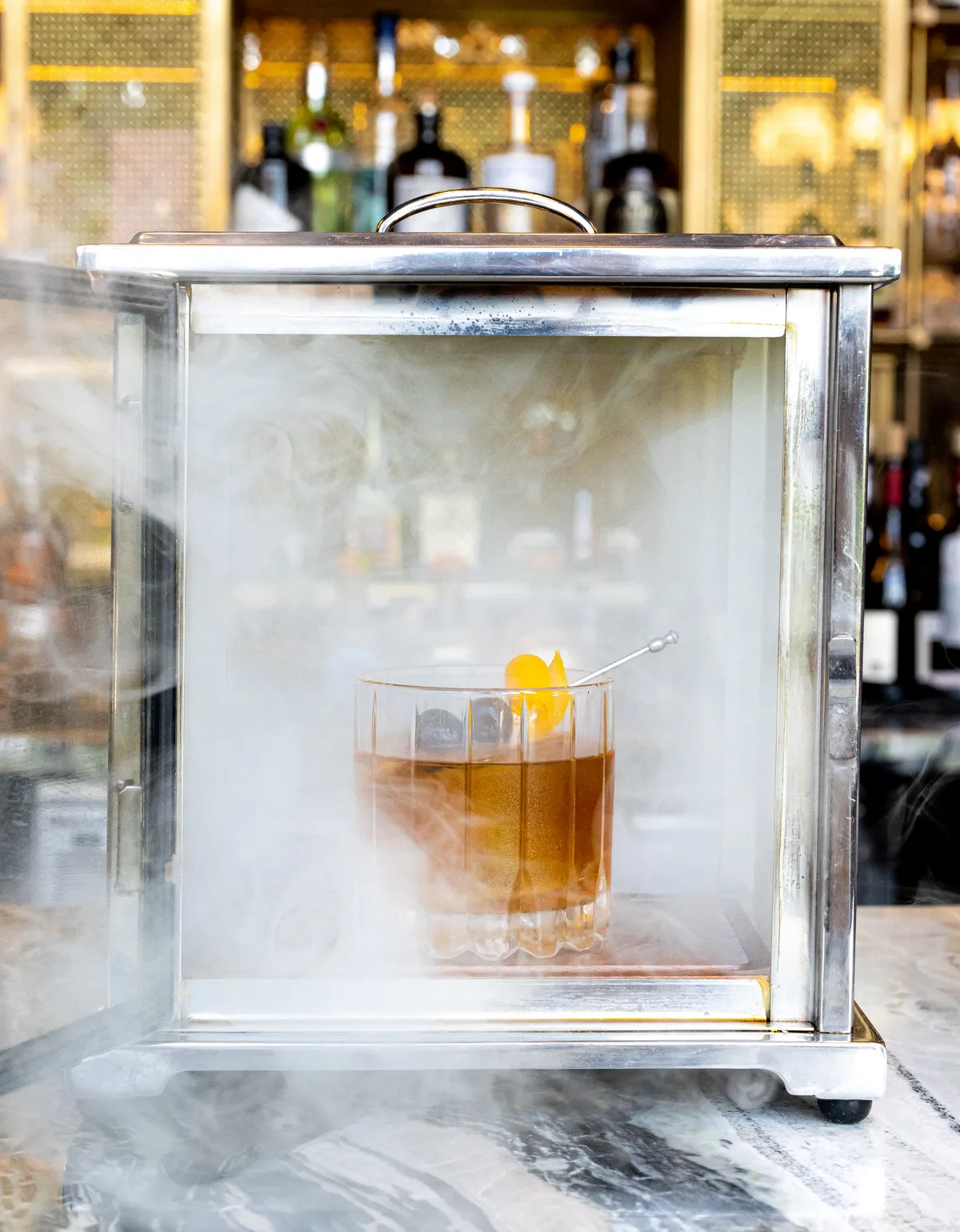 A Smoked Old Fashioned at Circle Bar. A cocktail in a steel glass case with smoke bellowing out. 