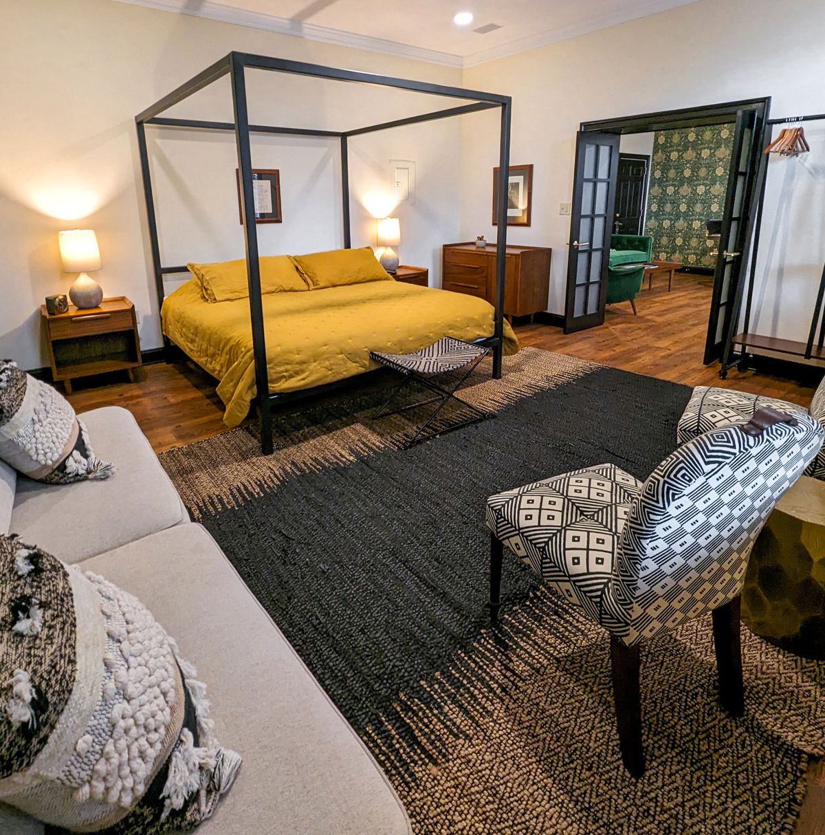 The king bedroom area in the Lincoln suite at the Union Hotel. The room has hardwood floors, a canopy bed with yellow and black patterned bedding, colorful throw pillows, and a seating area with armchairs and ottomans in various prints. French doors lead to an adjoining room.