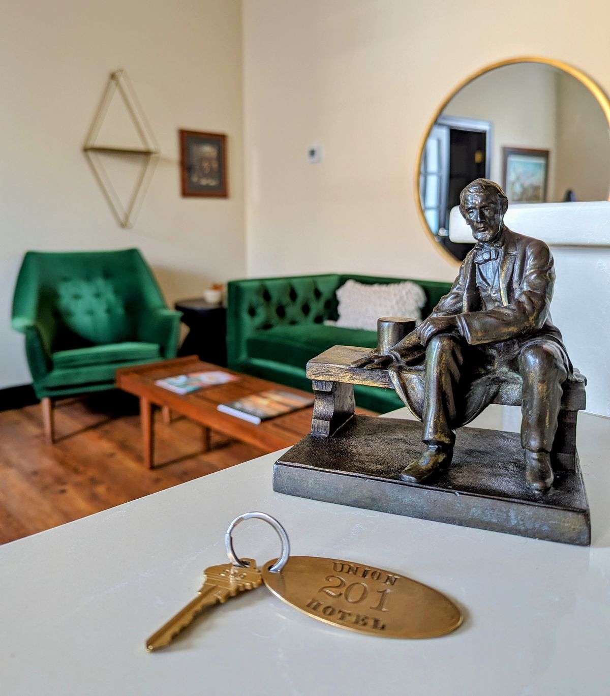 a closer look inside a room at the Union Hotel. A small bronze statue of Abe Lincoln is the focal point, with a room key labeled "Union 201 Hotel" in the foreground. Green velvet armchairs and a small wooden table can be seen in the background, giving the space an elegant, classic feel.