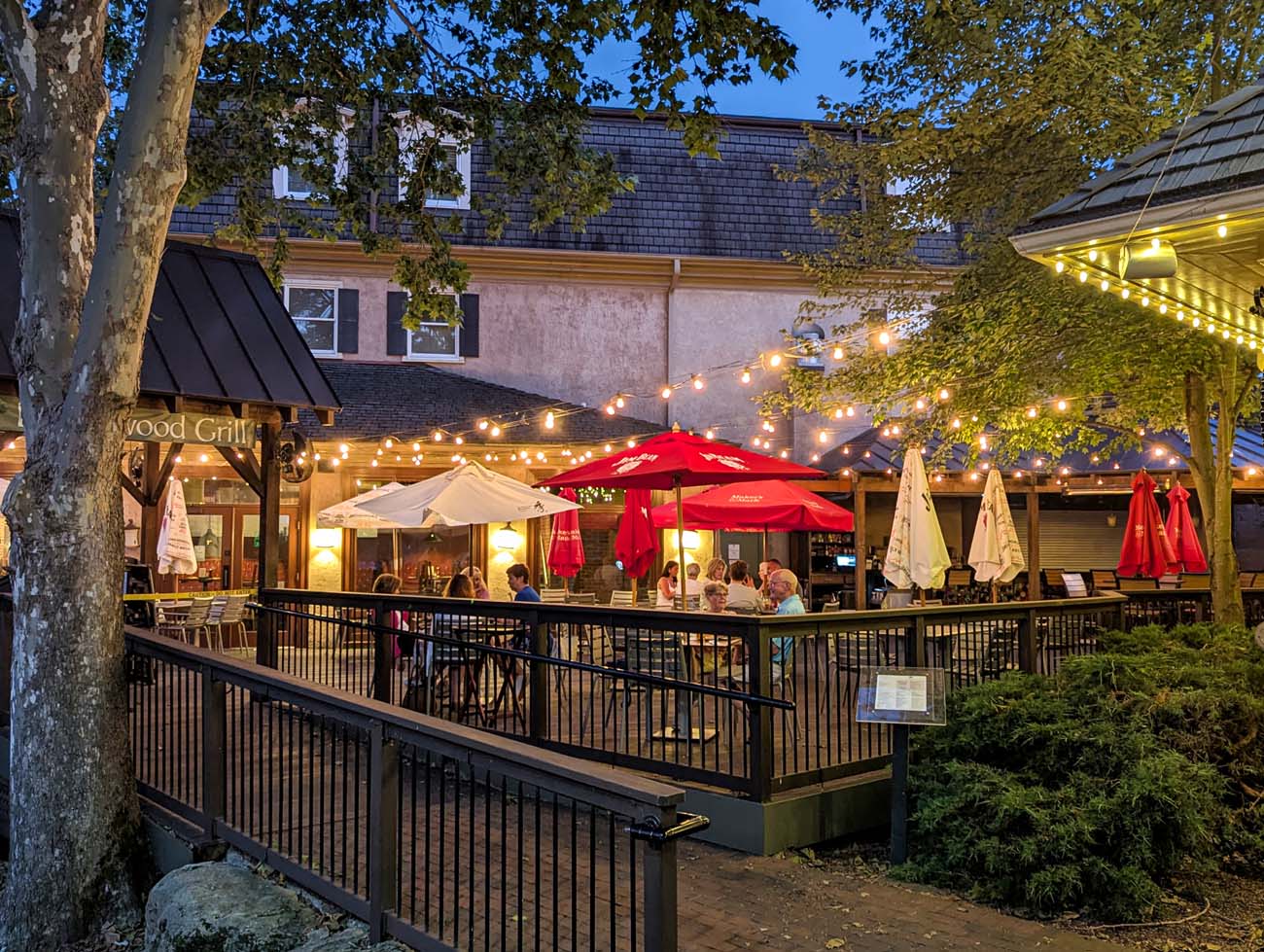 Evening view of Buttonwood Grill's outdoor dining patio with string lights overhead, red umbrellas, diners seated at tables, and colonial-style buildings in the background at Peddler's Village