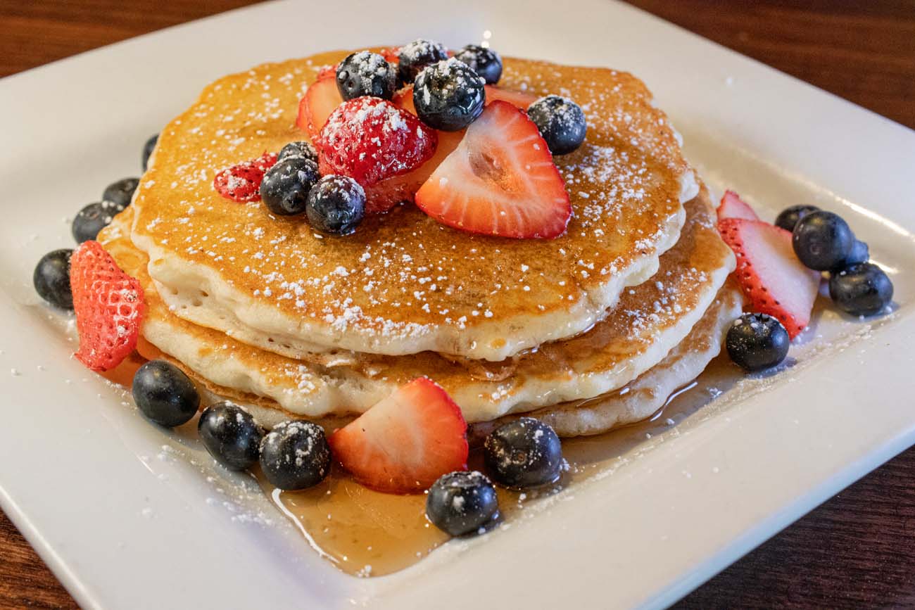 Stack of golden pancakes topped with fresh strawberries, blueberries, raspberries, powdered sugar, and maple syrup being poured from above, served on a white plate at Buttonwood Grill