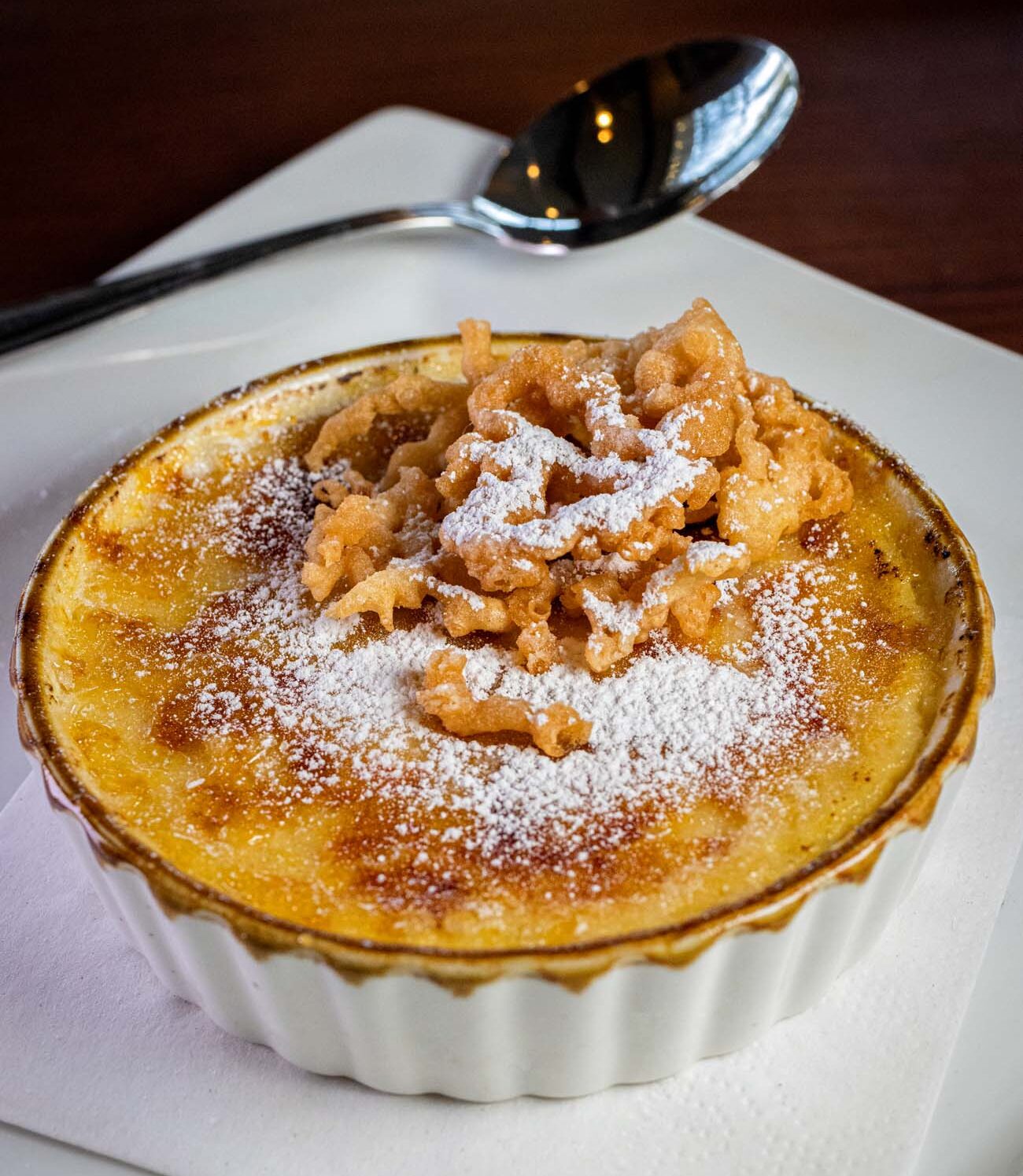 Layered funnel cake crème brûlée dessert served in a glass with whipped cream, crumble pieces, and caramelized sugar cubes on top, being eaten with a spoon at Cock N Bull