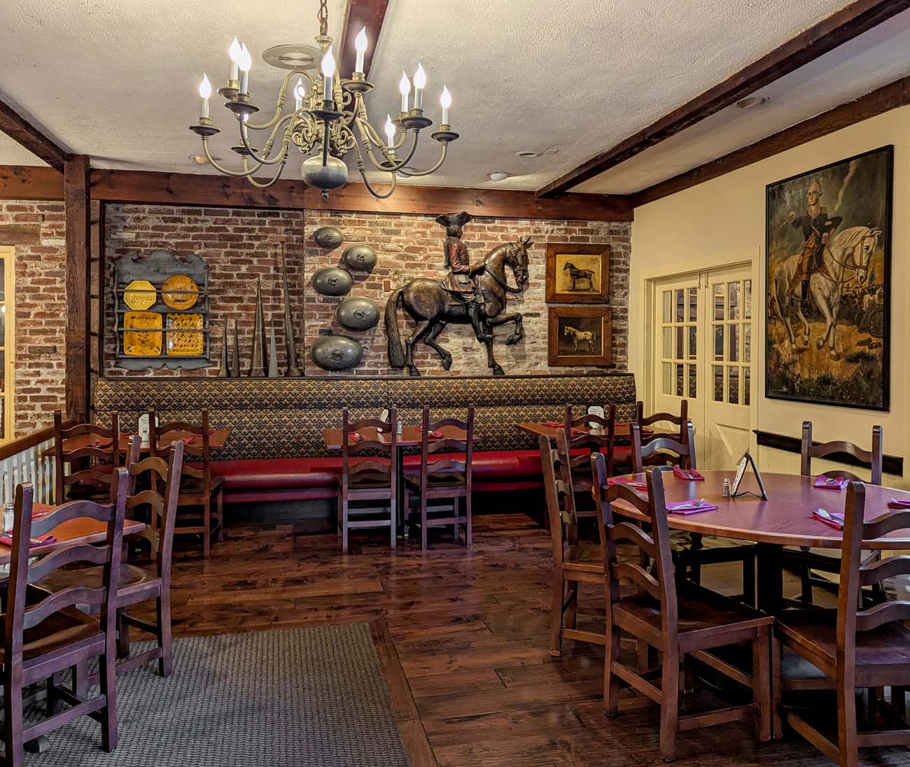 Interior of Cock 'n Bull restaurant showing colonial decor with exposed brick walls, wooden tables and chairs, brass chandelier, equestrian artwork, and traditional Pennsylvania country atmosphere