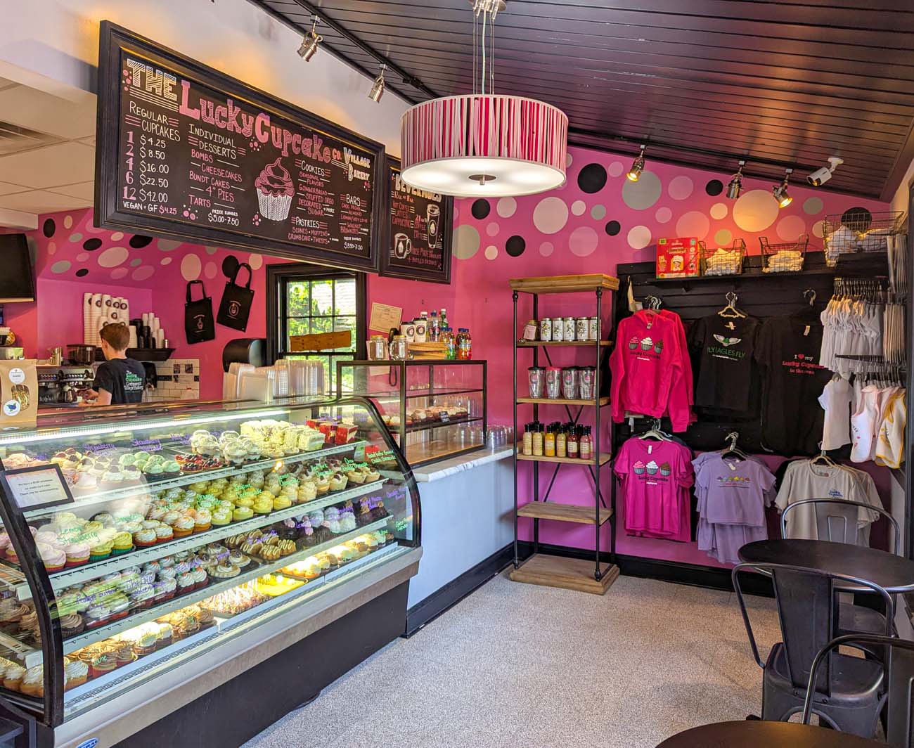 Interior of The Lucky Cupcake Company showing pink polka dot walls, curved glass display case filled with colorful cupcakes, menu boards, merchandise, and modern bakery lighting