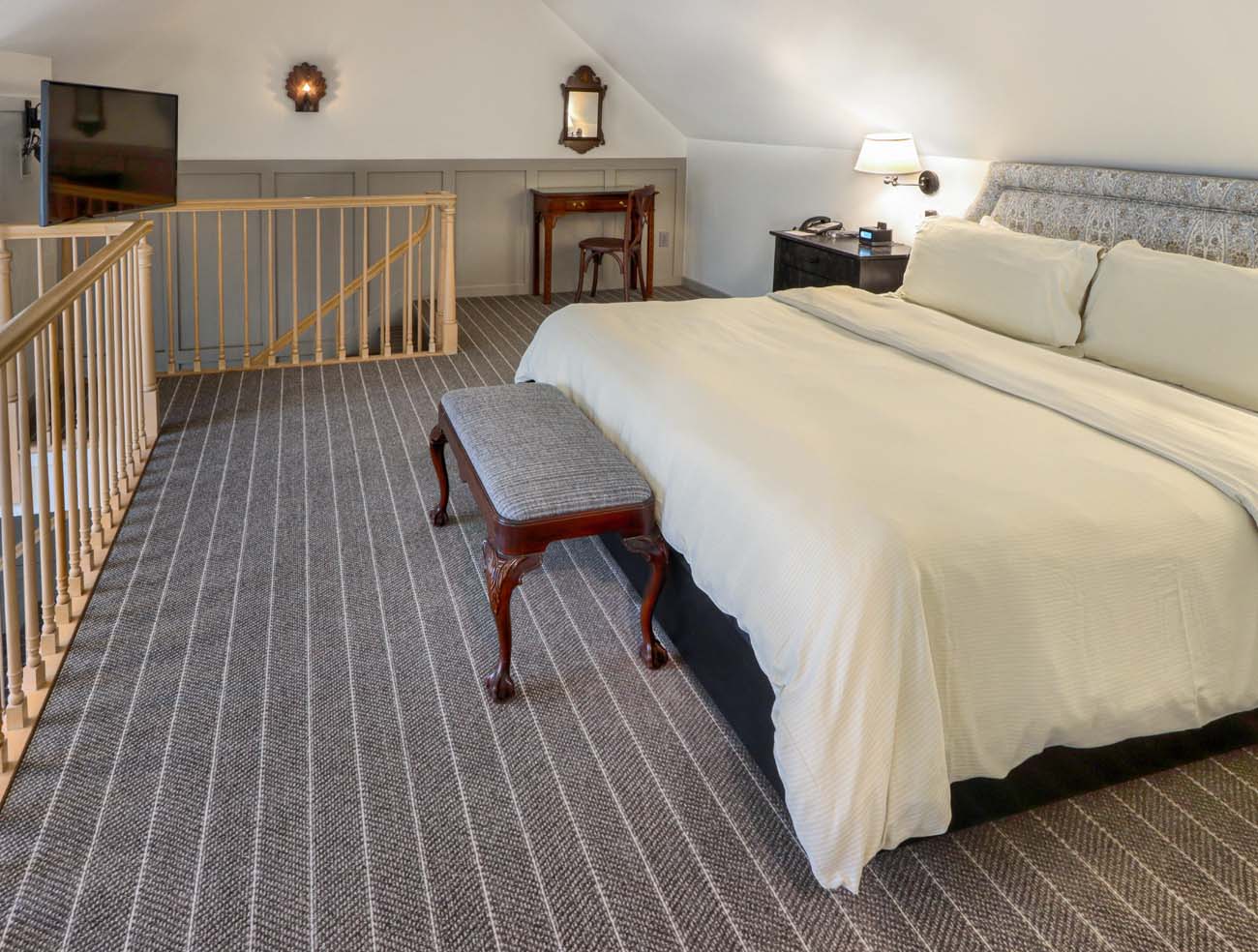 Upper level bedroom of Golden Plough Inn bi-level suite with king-size bed, white linens, wooden bench at foot of bed, wooden railing overlooking lower level, and traditional colonial-style furnishings