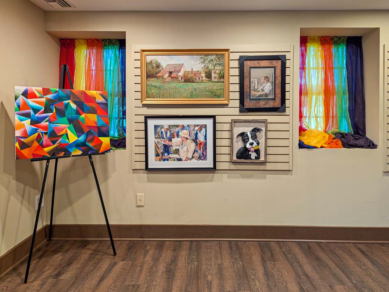 Art gallery display featuring colorful geometric painting on easel, framed artworks on slatwall display including landscapes and portraits, with rainbow fabric curtains framing windows during the Bucks Fever Art Exhibition