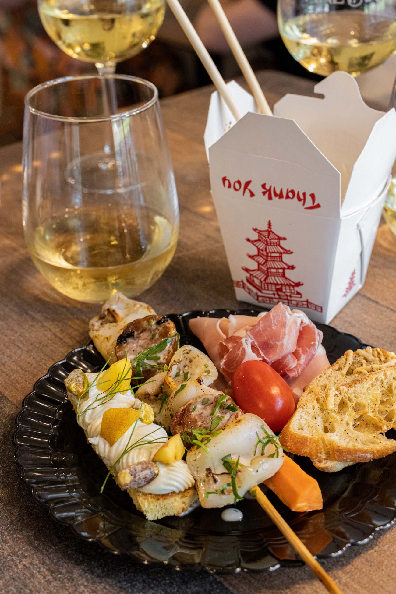 Elegant charcuterie plate with sliced meats, cheese, bread, and garnishes alongside white wine glasses and takeout container, photographed during Earl's New American Coast to Coast Wine Dinner