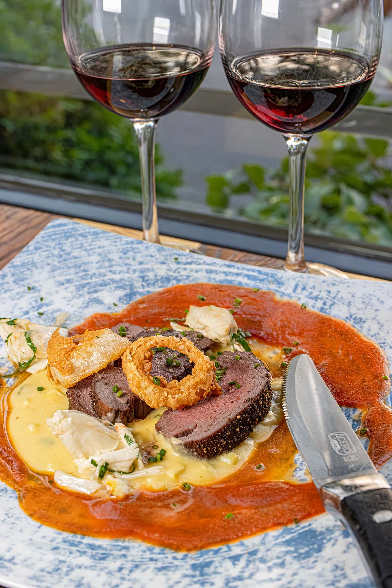 Perfectly sliced venison tenderloin with juniper berry crust, served over corn and poblano porridge with crispy onion garnish and red wine reduction sauce, paired with red wine glasses