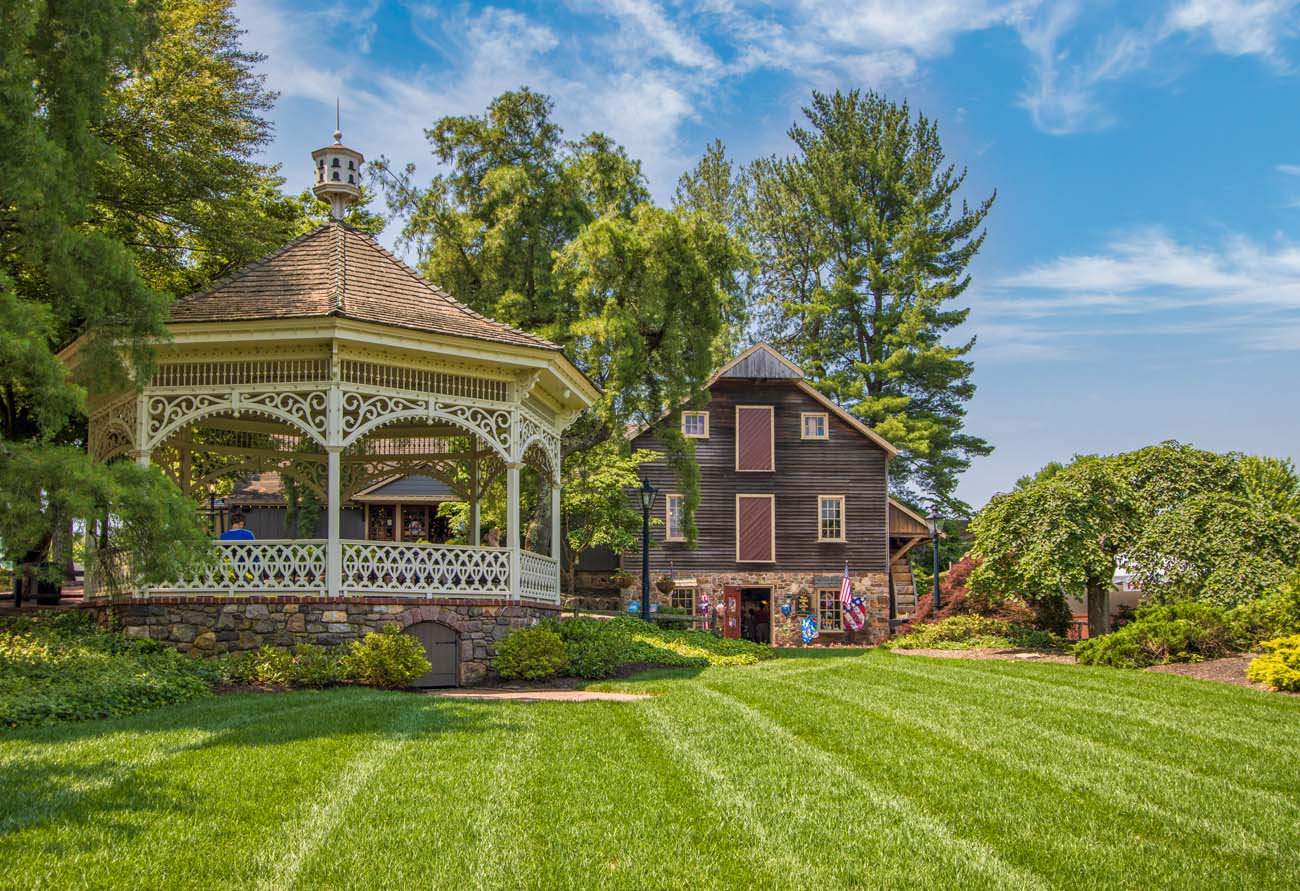 White Victorian gazebo with ornate trim on manicured lawn, surrounded by lush green trees and historic colonial buildings including a multi-story stone and wood structure in the background