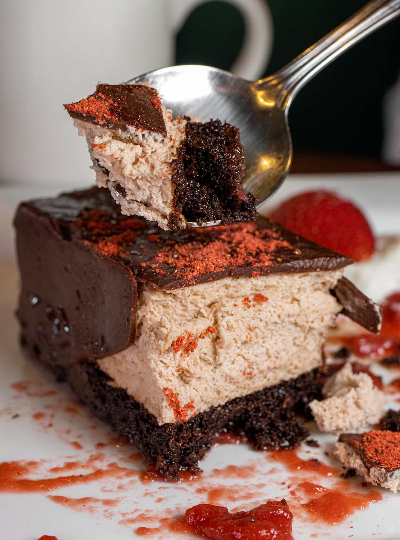 Elegant chocolate dessert with multiple layers including dark chocolate, strawberry mousse, and cake base, garnished with fresh strawberry sauce and being cut with fork to show interior layers