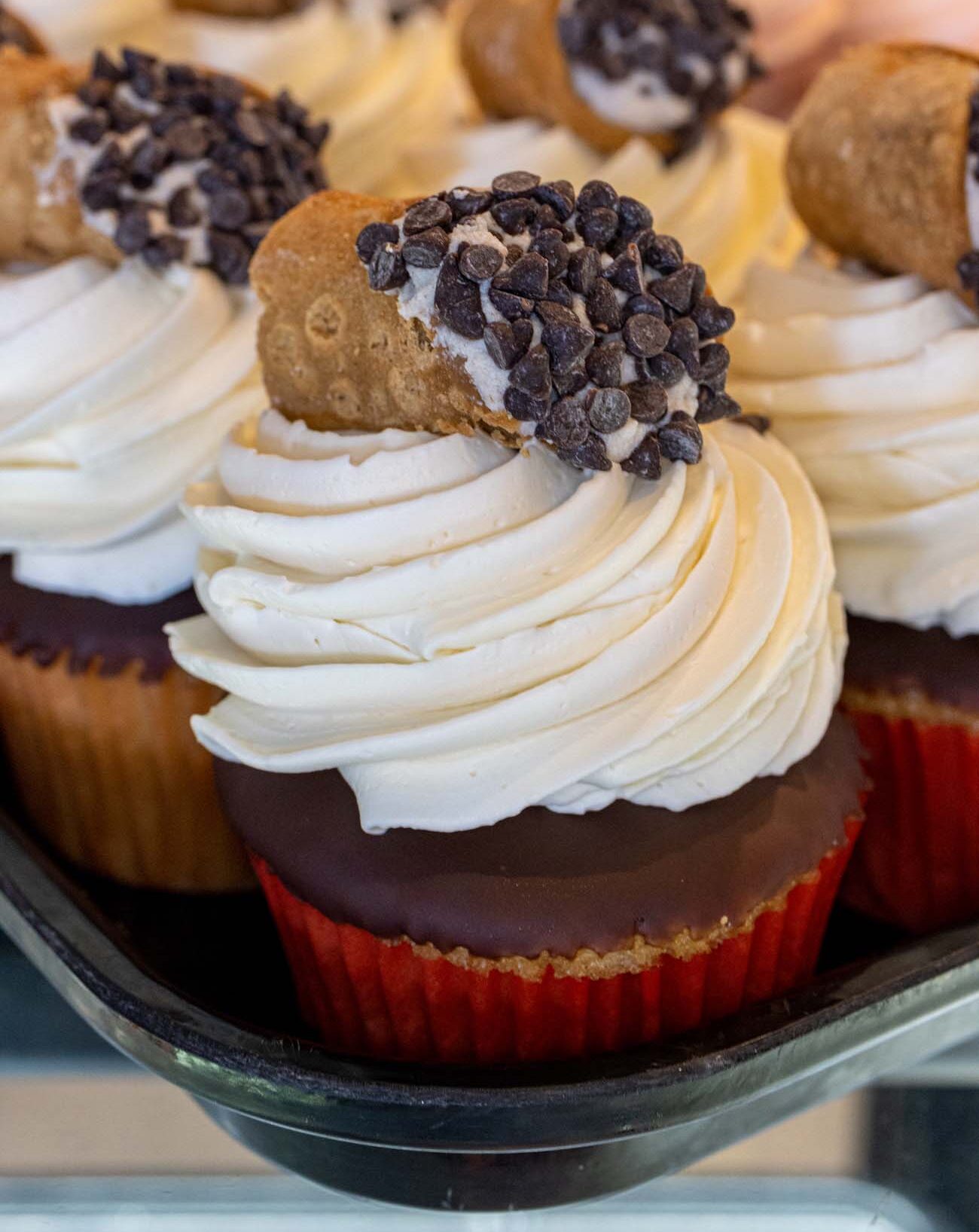 Display of gourmet cupcakes with chocolate bases, swirled vanilla buttercream frosting, mini chocolate chip cookies, and dark chocolate chips as garnish in a bakery display case