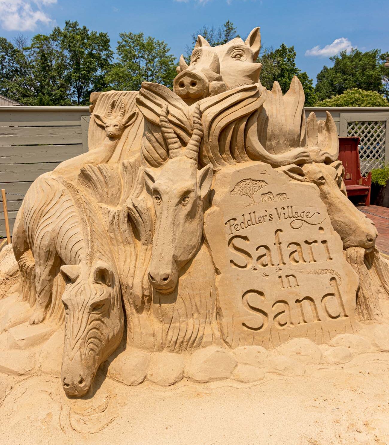 Large sand sculpture featuring multiple safari animals including lions, elephants, and zebras arranged around a central "Peddler's Village Safari in Sand" sign, displayed on brick walkway with colonial buildings and blue sky in background