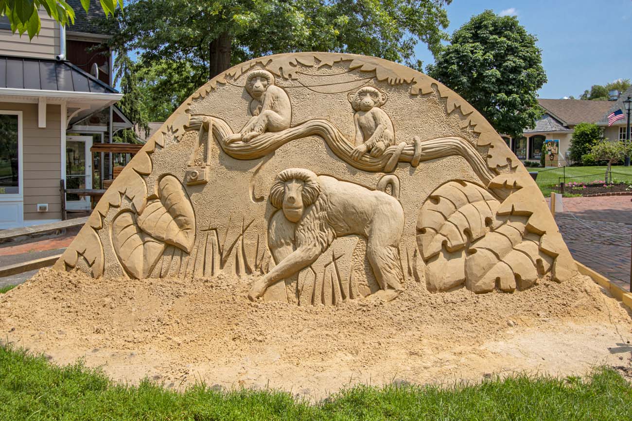 Circular sand sculpture featuring carved primates sitting on tree branches at the top surrounded by decorative leaf border, set on village green with colonial shops in background