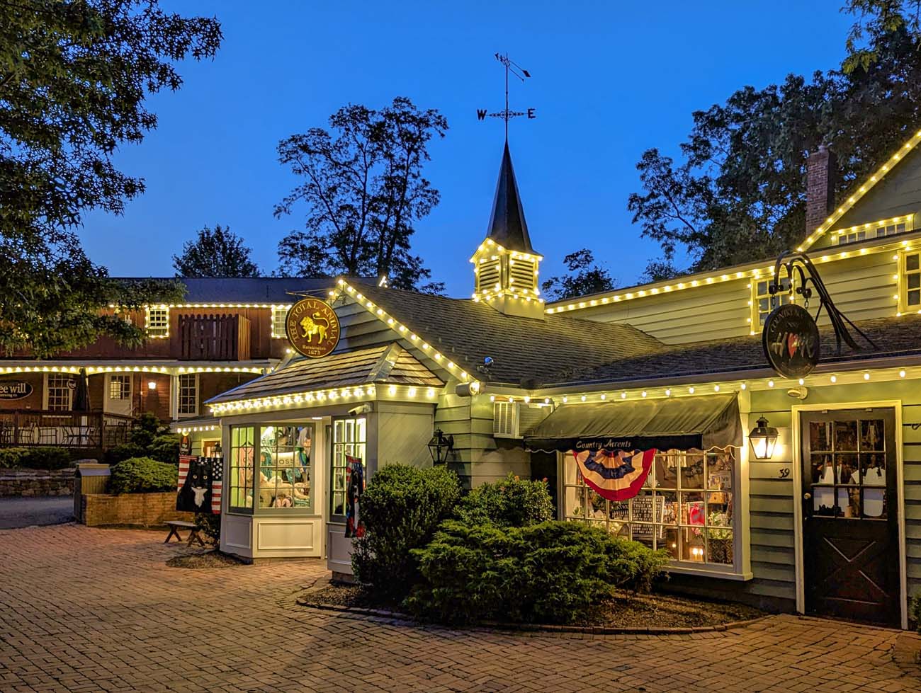 Evening view of illuminated colonial-style shops at Peddler's Village with string lights outlining building edges, Golden Plough Inn sign visible, American flag, and warm glow from shop windows against deep blue twilight sky