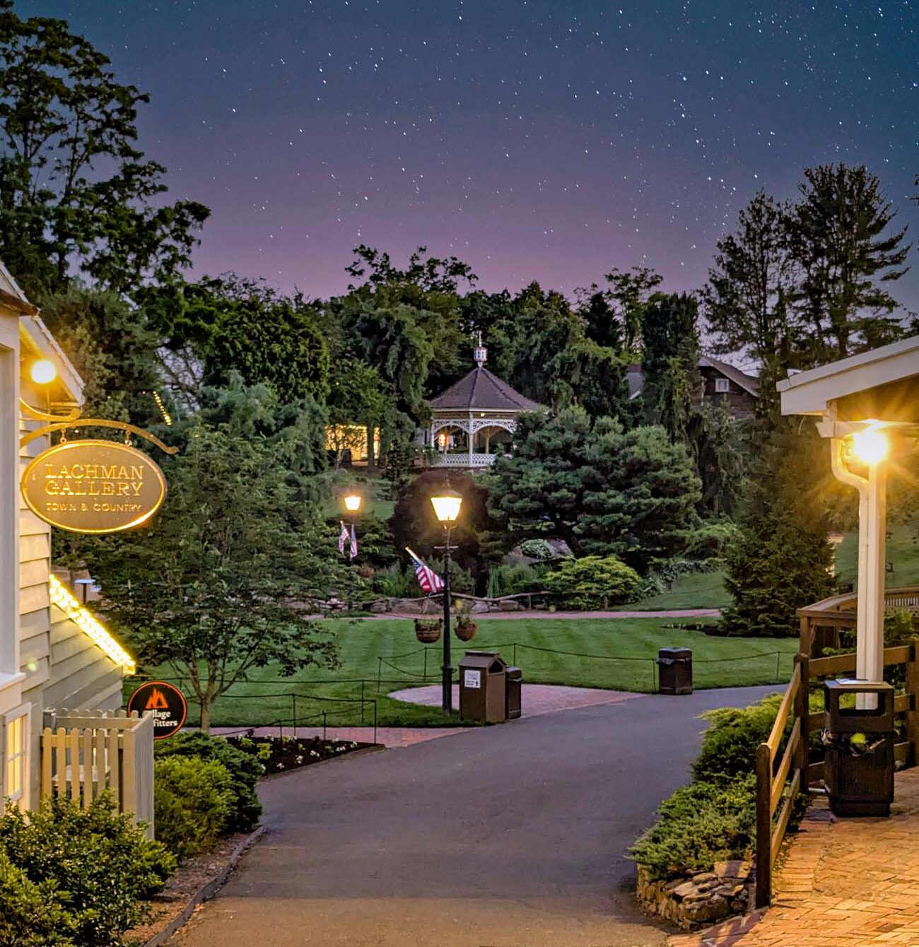 Magical nighttime scene of Peddler's Village showing starry sky with purple and blue hues, illuminated gazebo in background, lit pathways, manicured lawns, and shop signs including Lachman Gallery, creating enchanting village atmosphere