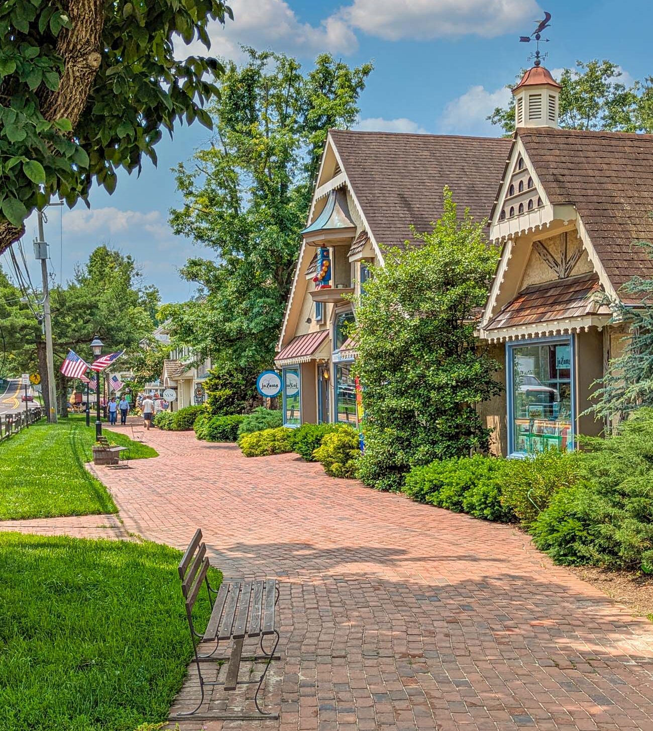 Charming brick walkway lined with Victorian-style shops featuring decorative gingerbread trim, American flags, manicured gardens, wooden bench, and visitors strolling through the tree-lined shopping district on sunny summer day