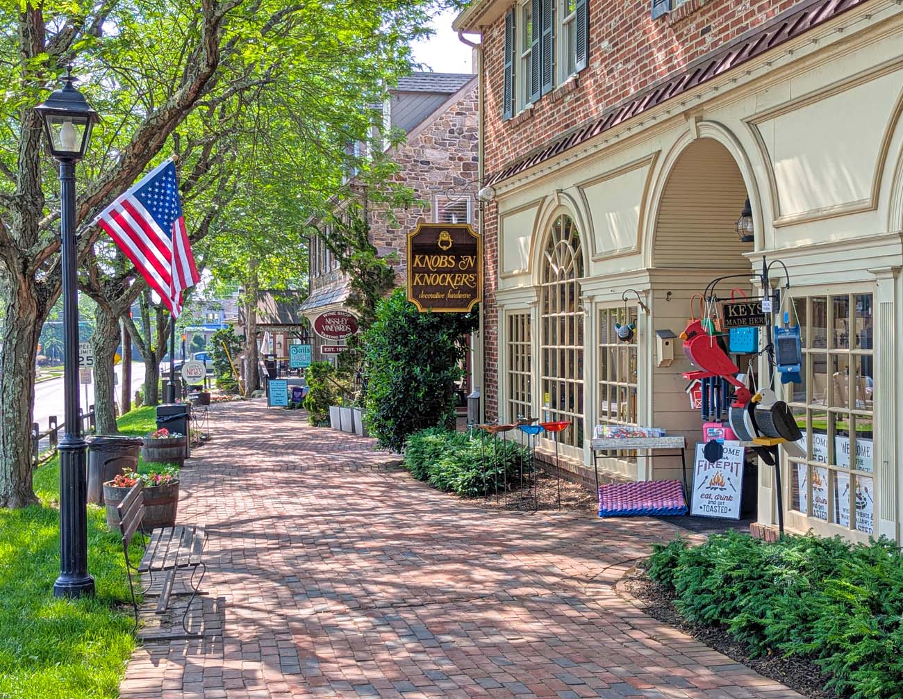 Tree-lined brick walkway through historic shopping district featuring colonial architecture with mix of brick and stone buildings, American flag, traditional lamp posts, shop signs including "Knobs and Knockers," and summer landscaping