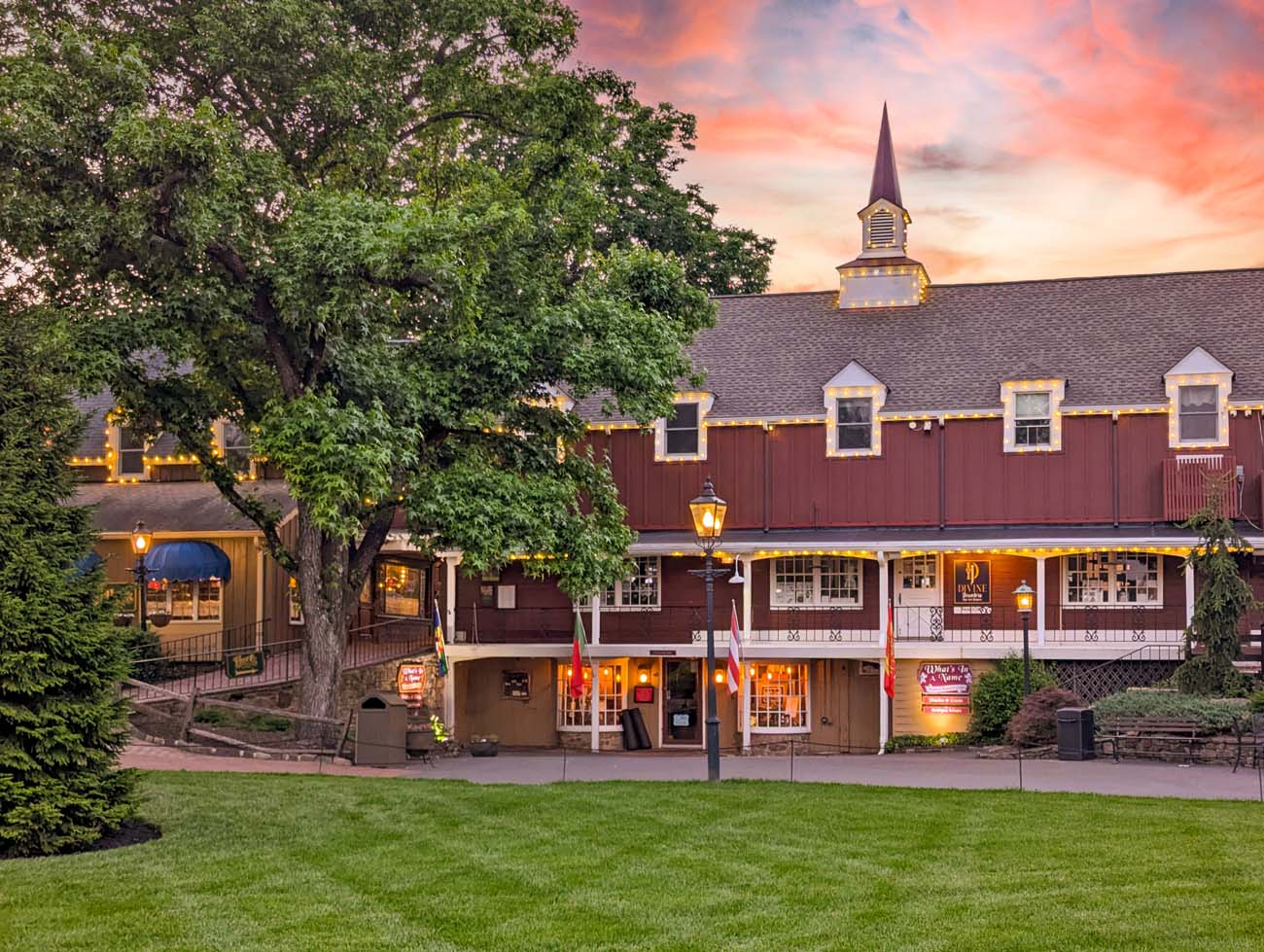 Dramatic sunset view of Peddler's Village featuring colonial buildings with red siding, church steeple, string lights, and warm window glow against spectacular orange and pink sunset sky with large mature trees silhouetted in foreground