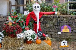 This image shows a festive Halloween display at Peddler's Village featuring a scarecrow-like figure dressed as a skeleton wearing a red Santa Claus outfit. The figure is posed with one arm outstretched, holding a wrapped present. It stands among hay bales, pumpkins, and potted flowers, including red and white chrysanthemums. Several Halloween-themed signs are placed around the scene, including one that reads "Hey bootiful," another that says "Trick or Treat," and a "Happy Halloween" sign with a cartoon ghost and pumpkin. The background features a rustic stone wall and greenery, adding to the seasonal charm.