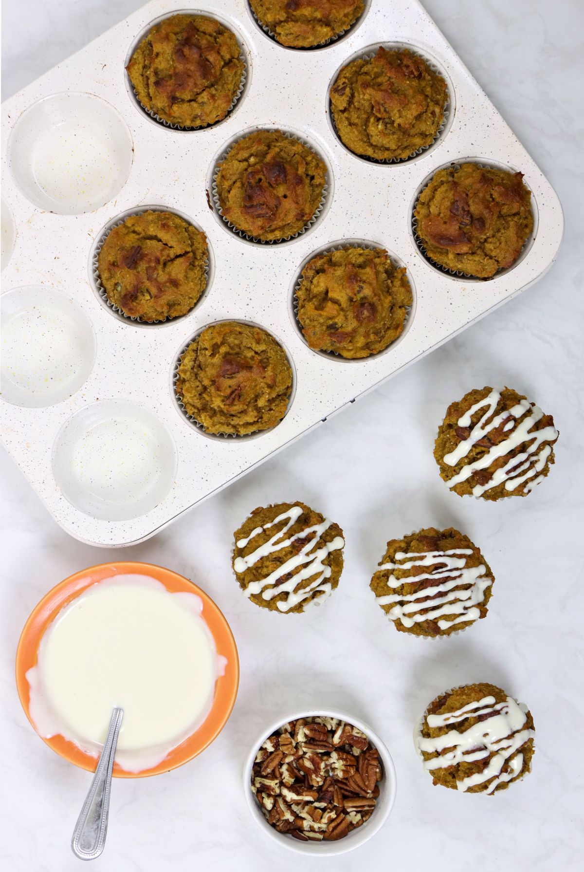 White muffin tins contain freshly baked pumpkin pecan muffins, some decorated with a drizzle of white icing. A small bowl of chopped pecans and a bowl of oranges with icing visible in the corner.