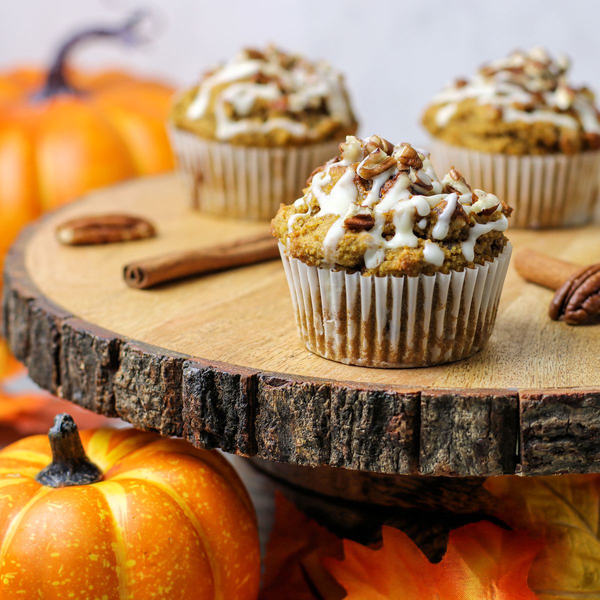 Delicious pumpkin pecan muffins topped with cream cheese glaze and chopped pecans displayed on a rustic piece of wood. Decorative mini pumpkins and fallen leaves surround the muffins to create a cozy fall atmosphere.