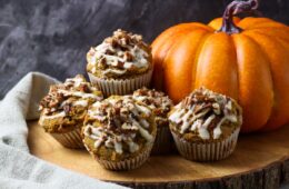 Four golden-brown pumpkin pecan muffins with a sweet glaze and pecan topping, placed on a wooden board with a large pumpkin in the background. A perfect autumn snack or dessert.