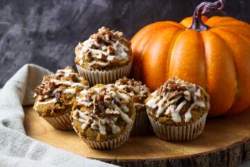 Four golden-brown pumpkin pecan muffins with a sweet glaze and pecan topping, placed on a wooden board with a large pumpkin in the background. A perfect autumn snack or dessert.