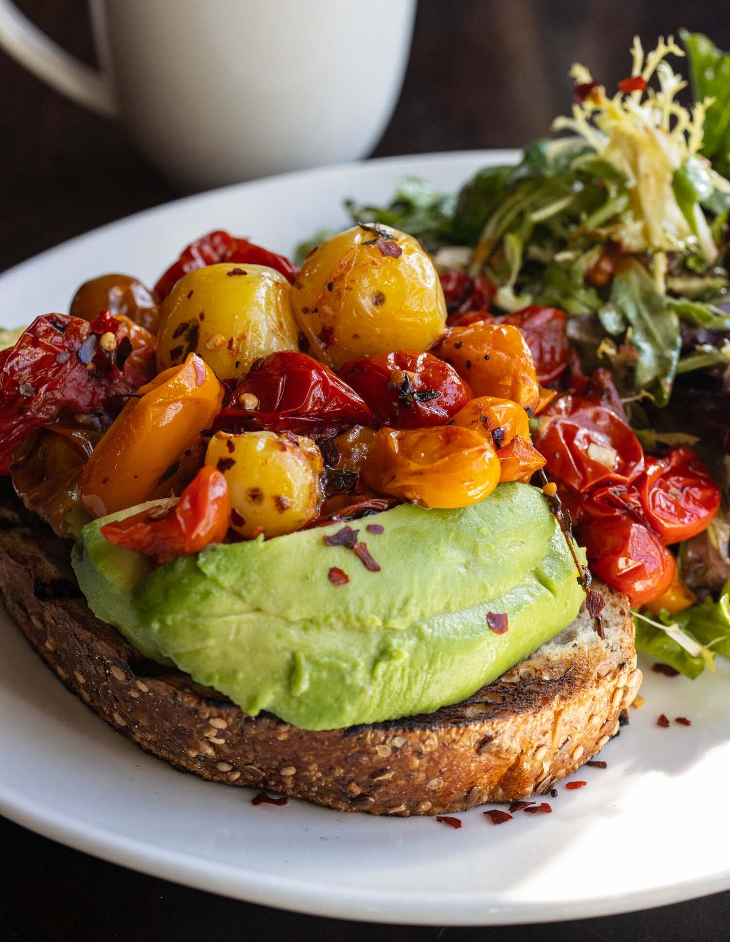 Avocado toast on ancient grain bread topped with roasted colorful cherry tomatoes and chili flakes, served with a side salad and coffee