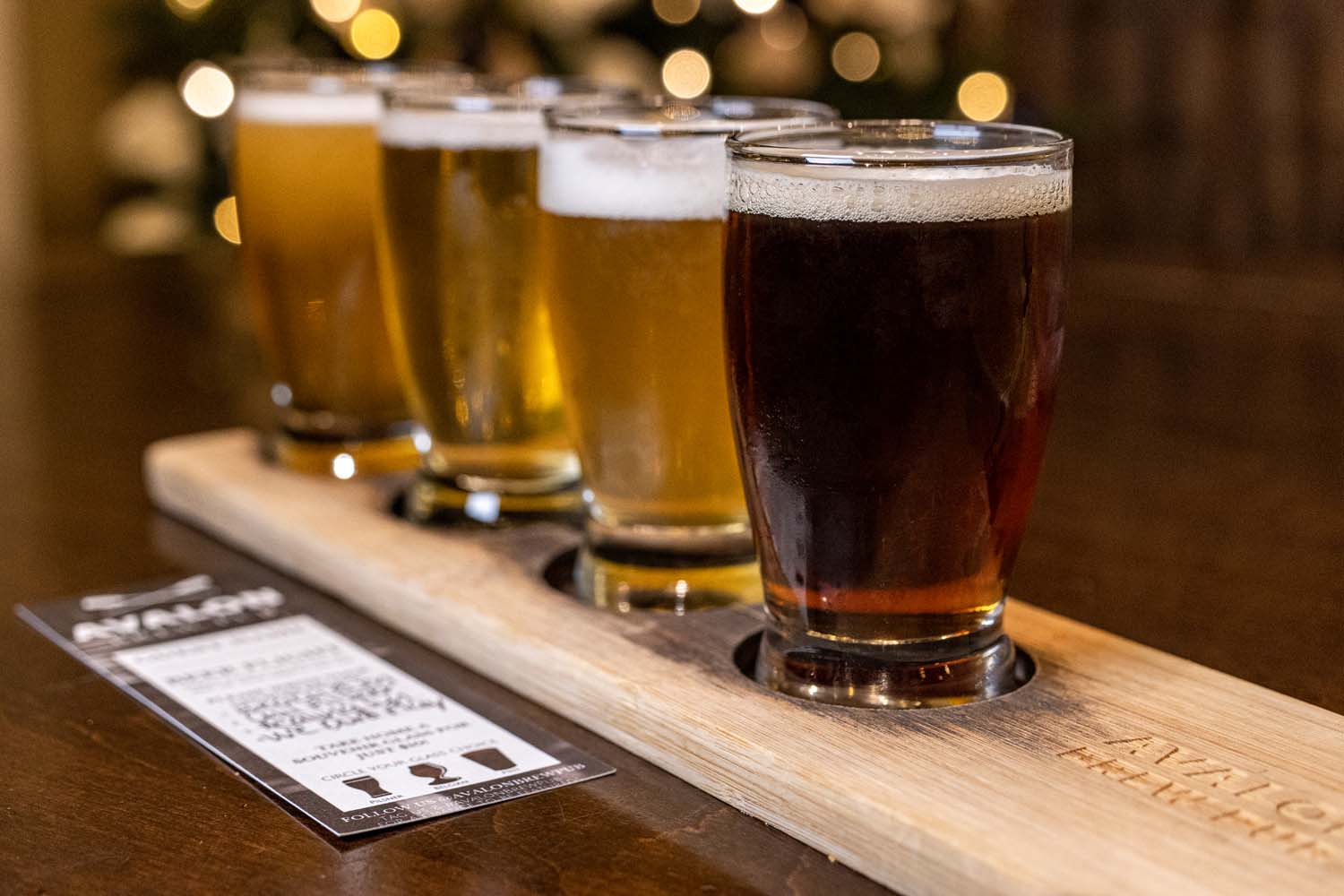 A wooden flight board with four beer samples ranging from light golden to dark amber, with a tasting menu card