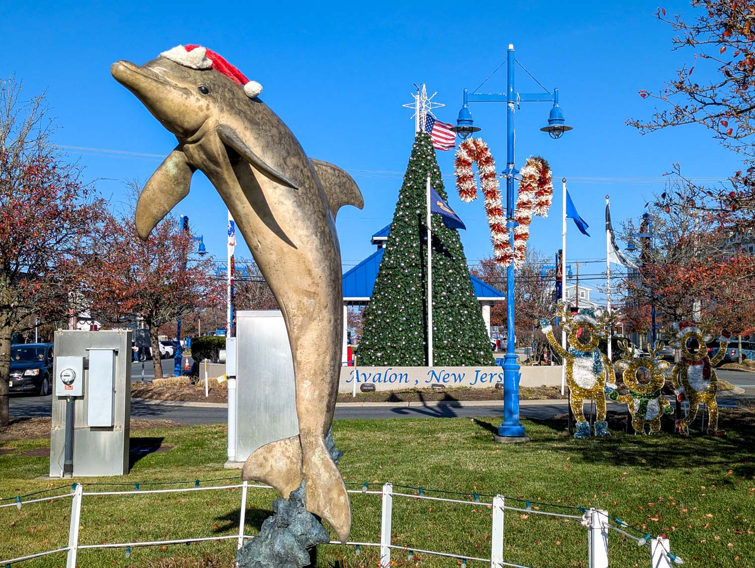 Large bronze dolphin statue wearing a Santa hat in front of a lit Christmas tree and light-up reindeer decorations in Avalon's town center, with blue lampposts and American flag visible against a bright winter sky