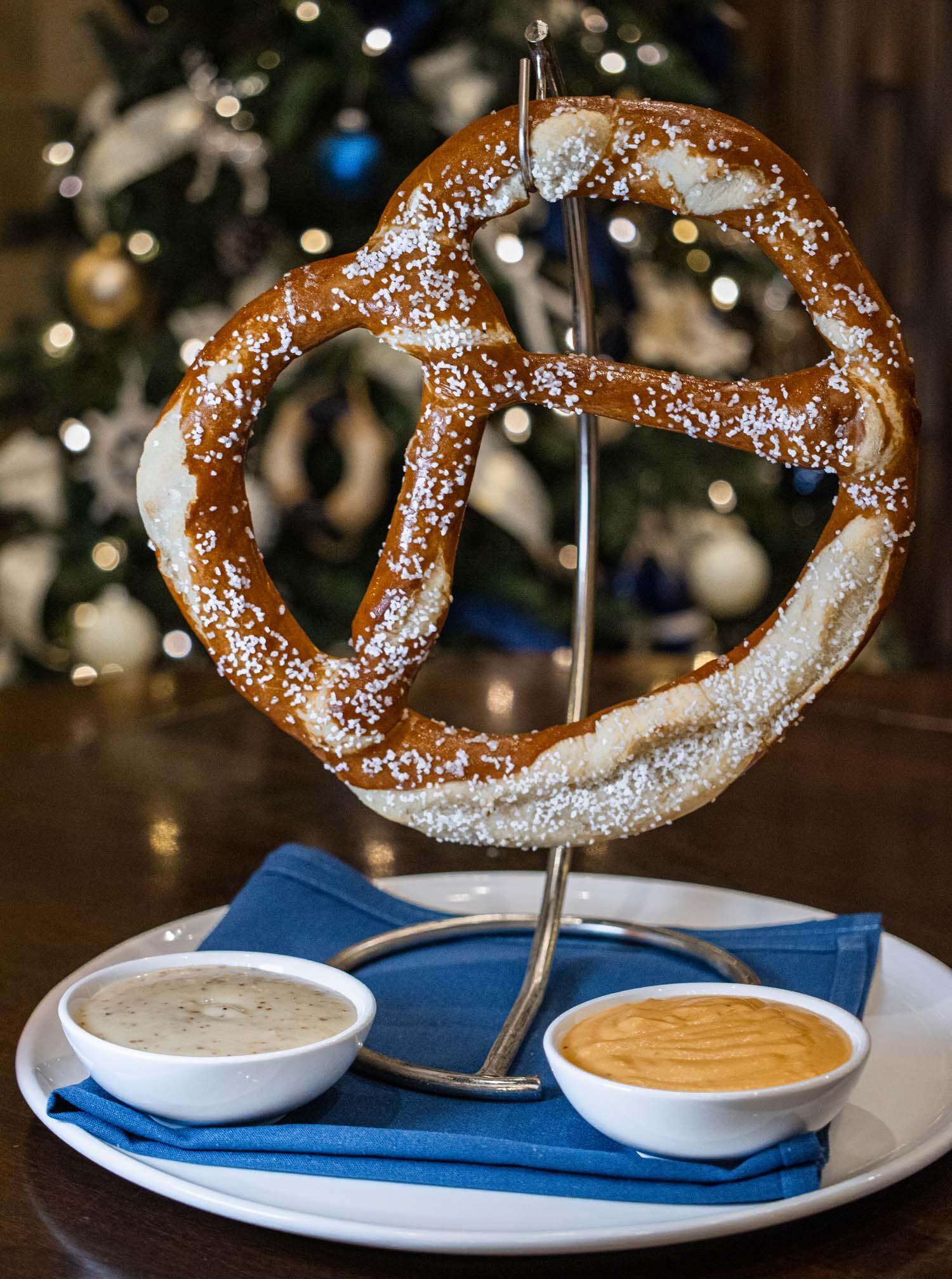 Giant Bavarian pretzel displayed vertically on a metal stand, dusted with coarse salt against a backdrop of twinkling Christmas tree lights, served with beer cheese and grain mustard aioli in white ceramic bowls on a navy blue napkin