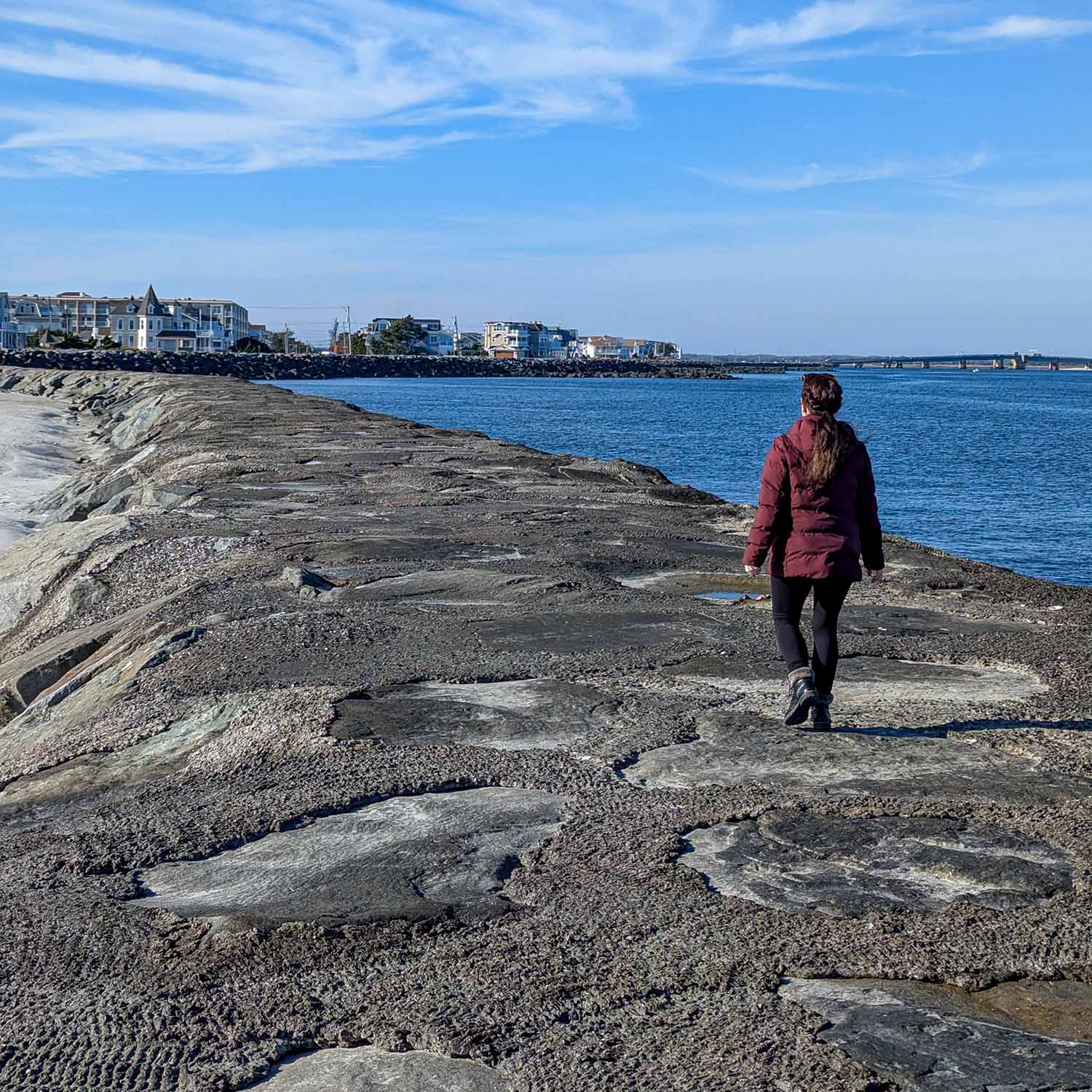 Person walking along the rocky Avalon Seawatch jetty on a clear winter day, with coastal homes and the blue Atlantic Ocean visible in the background