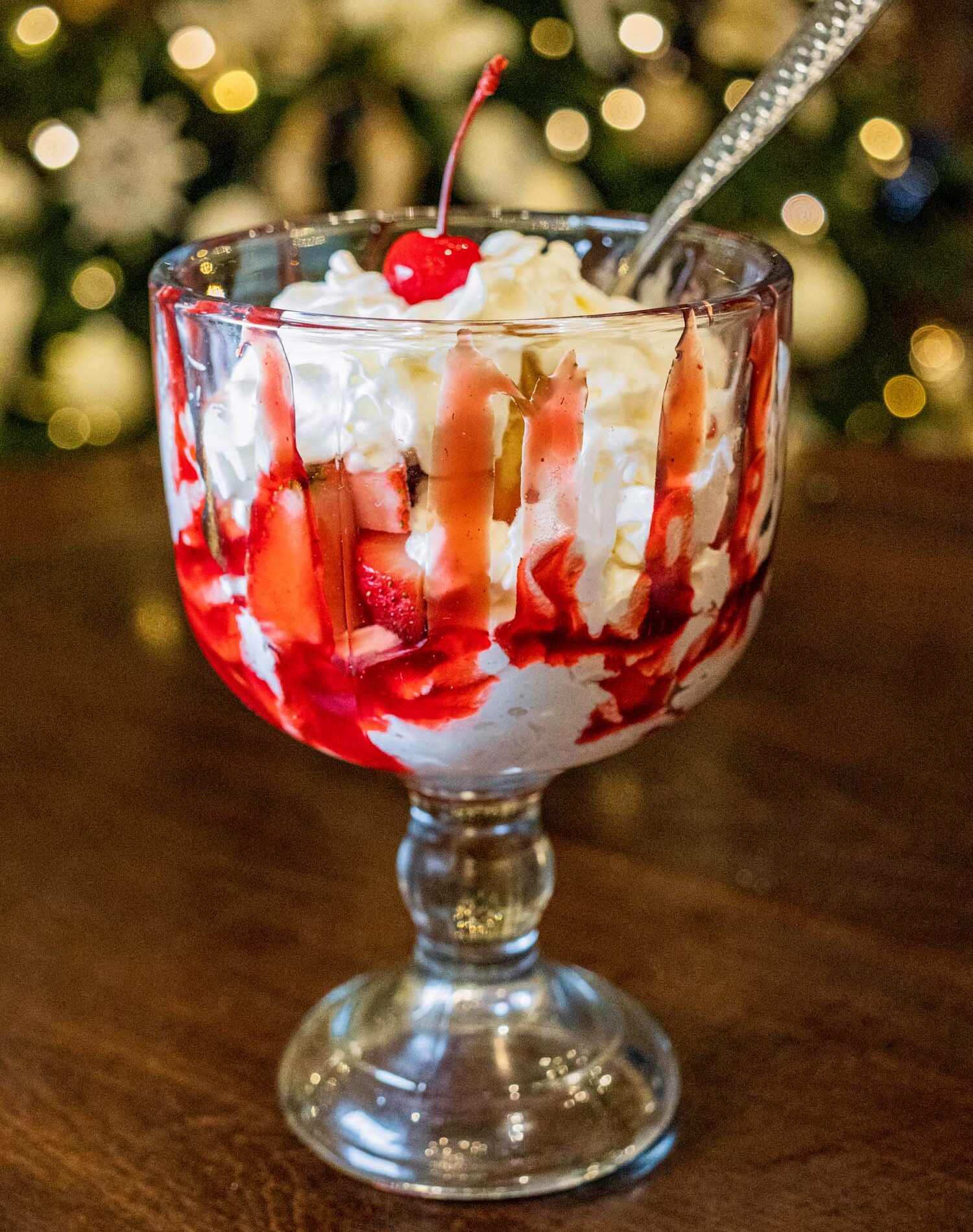 Layered cheesecake dessert served in an elegant glass goblet featuring whipped cream, fresh strawberries, and graham cracker crumbs, topped with a maraschino cherry, photographed against soft holiday lights