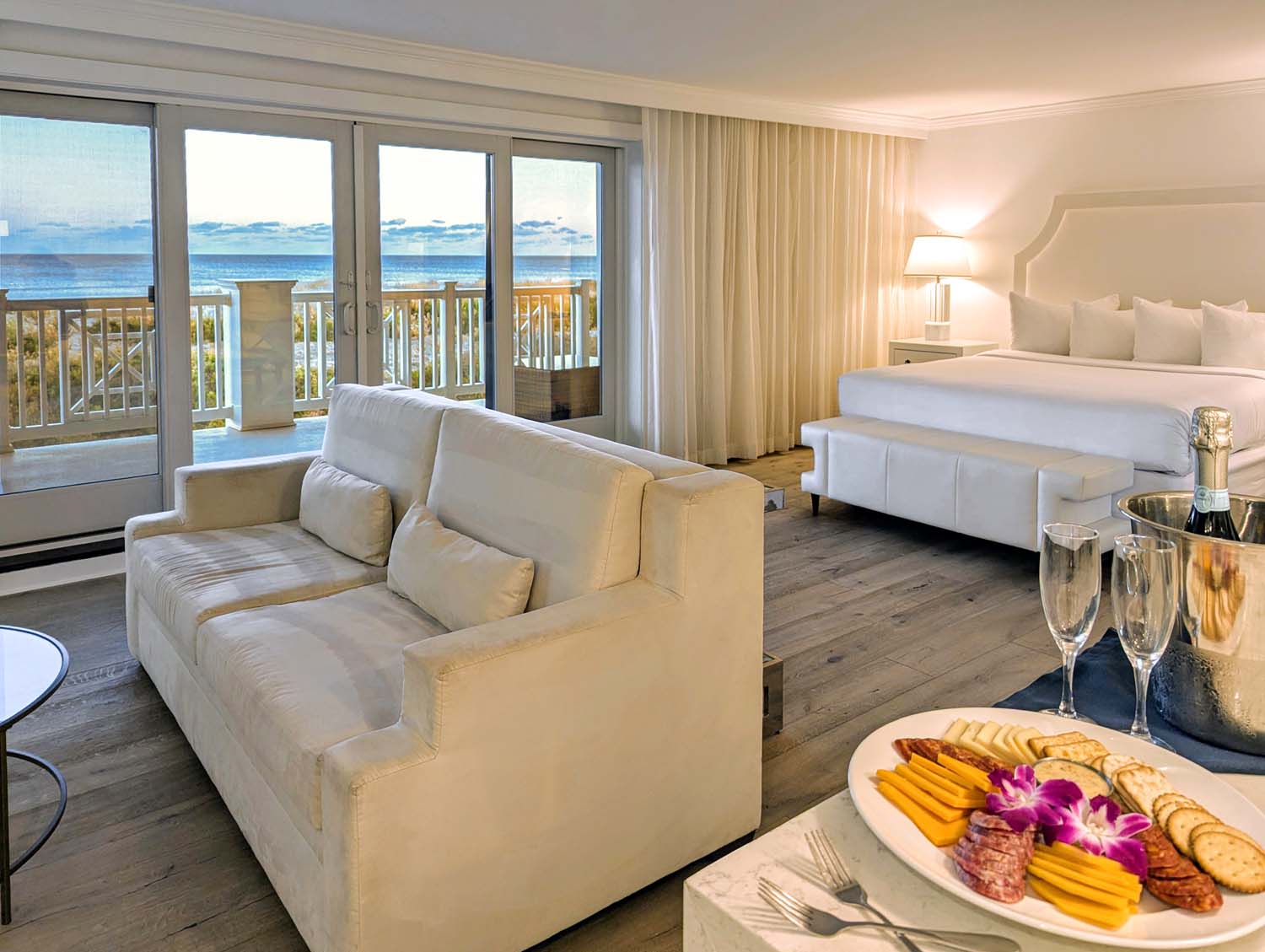 Luxurious oceanfront studio featuring a white king bed, matching sleeper sofa, and floor-to-ceiling windows opening to a balcony with Atlantic Ocean views. A charcuterie board with meats, cheeses, and a purple orchid garnish sits beside champagne flutes on a marble side table