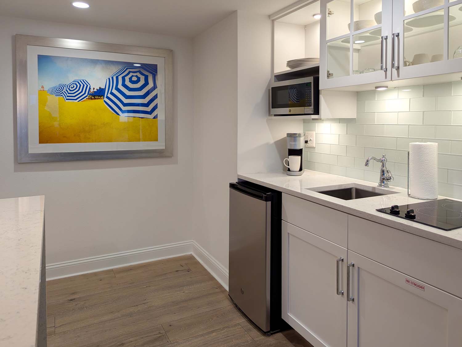 Modern white galley kitchen with beachy artwork featuring blue and white striped umbrellas, stainless steel appliances, subway tile backsplash, Keurig coffee maker, and quartz countertops with coordinated light wood flooring