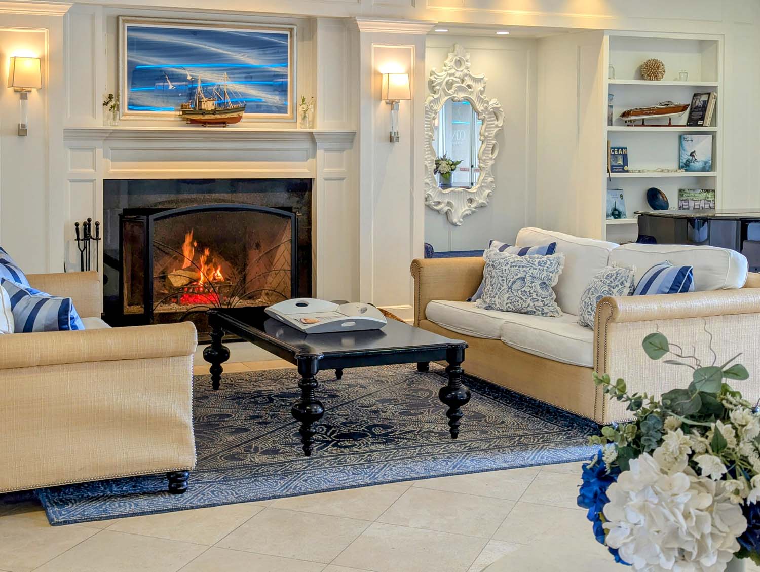 Elegant coastal-style lobby with roaring fireplace, nautical artwork and model ship above mantle, ornate white mirror, cream upholstered sofas, black coffee table, and blue patterned rug, accented with white hydrangea bouquet