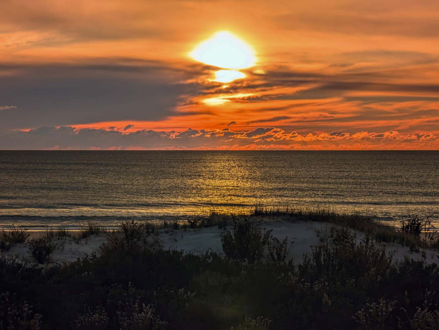 Dramatic winter sunrise over the Atlantic Ocean, with orange and gold clouds illuminated by the rising sun, casting golden reflections on the water and silhouetting the dune grass in the foreground