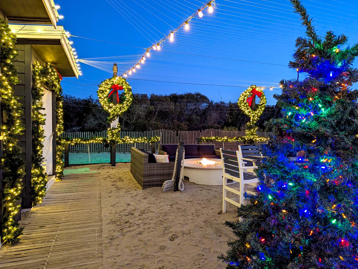 Beachside lounge area at dusk with illuminated Christmas wreaths with red bows, string lights overhead, boardwalk path, and a colorfully lit Christmas tree beside fire pit seating