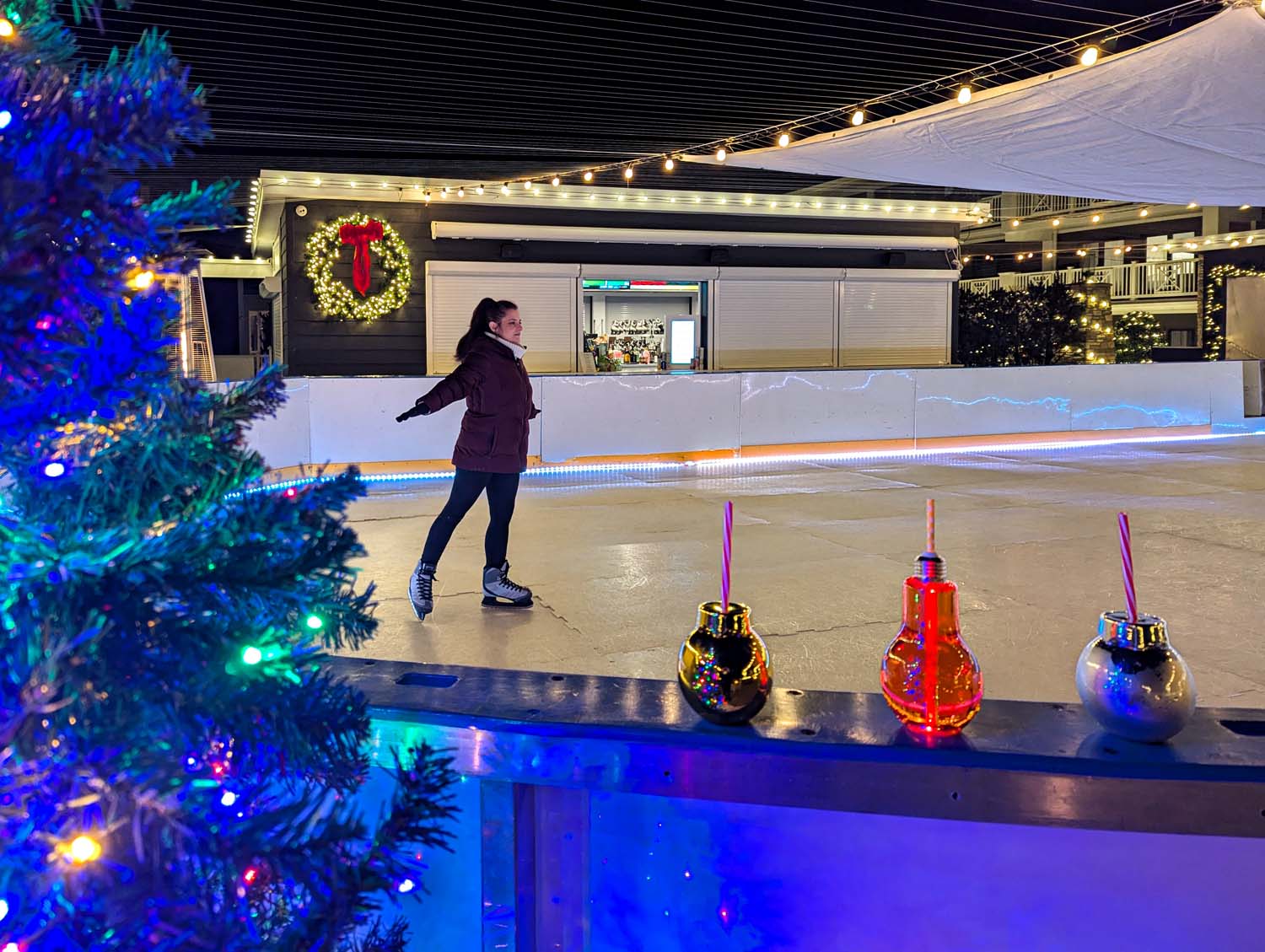 A nighttime view of the ICONA Winter Wonderland skating rink with LED lighting, holiday wreaths, and festive ornament-shaped cocktails displayed along the rink's edge. A girl is skating and smiling. 