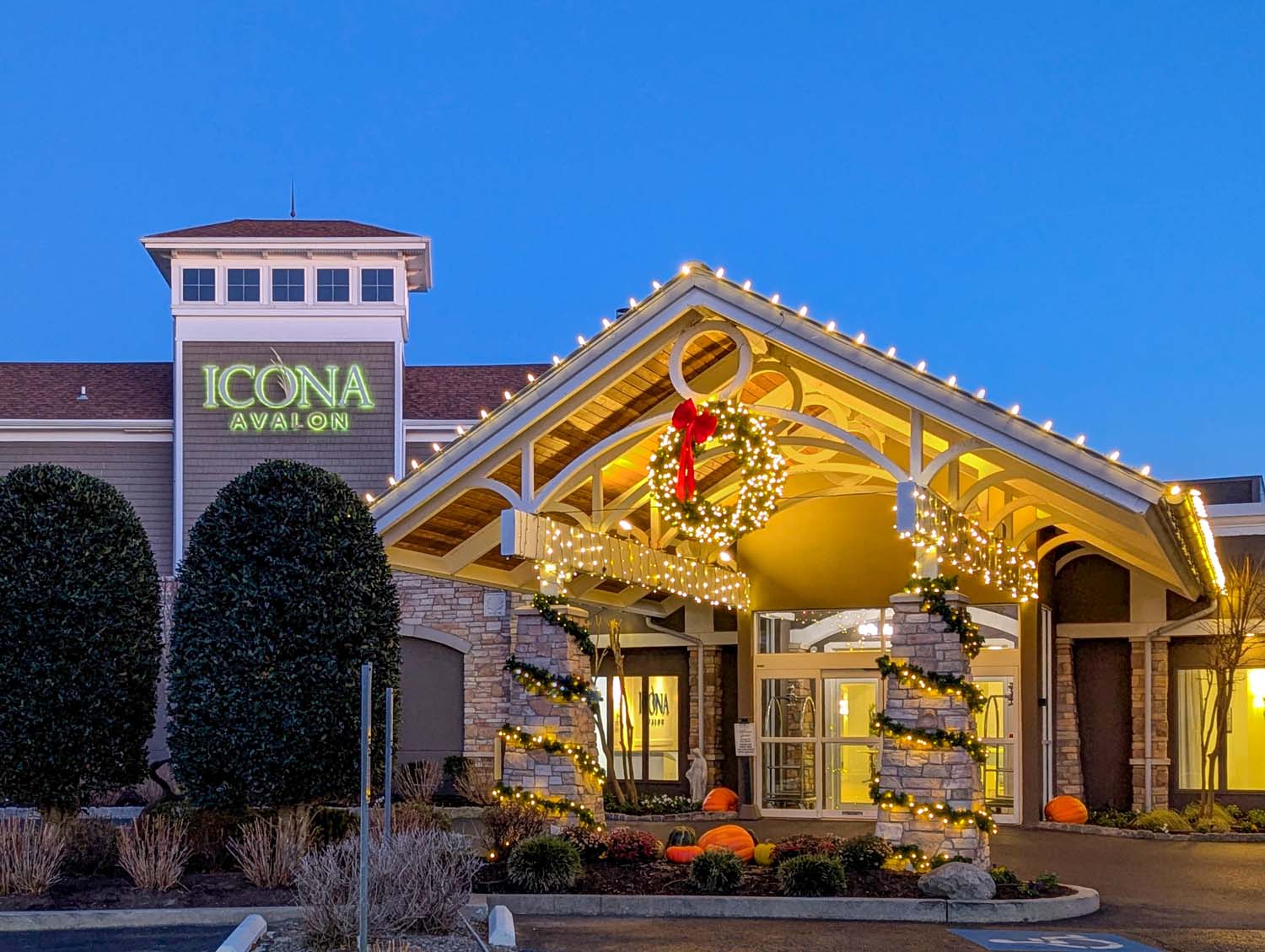 The ICONA Avalon entrance at dusk featuring a glowing green neon sign, stone facade, and holiday decorations including wreaths with red bows and white string lights